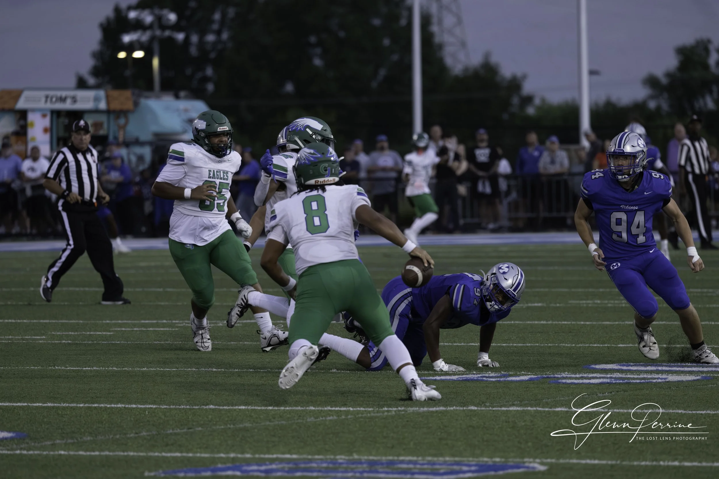 A football game in progress with players from the Eagles in white and green jerseys and the Vikings in blue jerseys. The Eagles player is holding the football and is about to be tackled by the Vikings player on the ground. Other players are running a