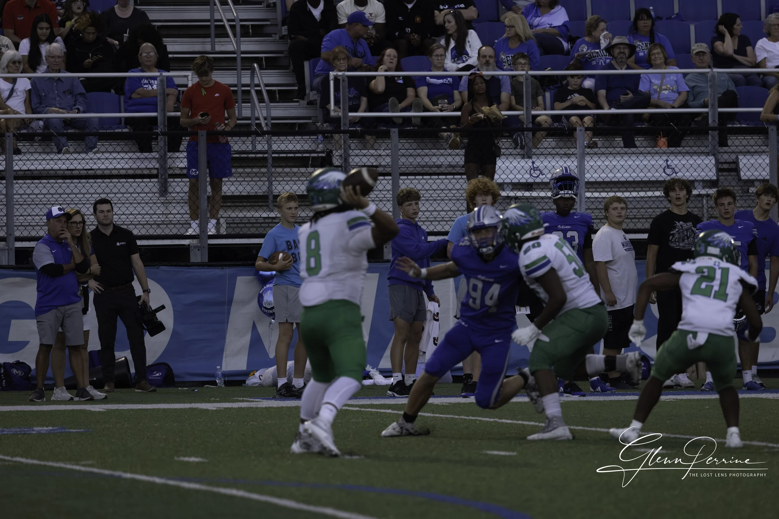 Football game with players in blue and green uniforms, some wearing helmets, on the field. Spectators seated in bleachers in the background watching the game.
