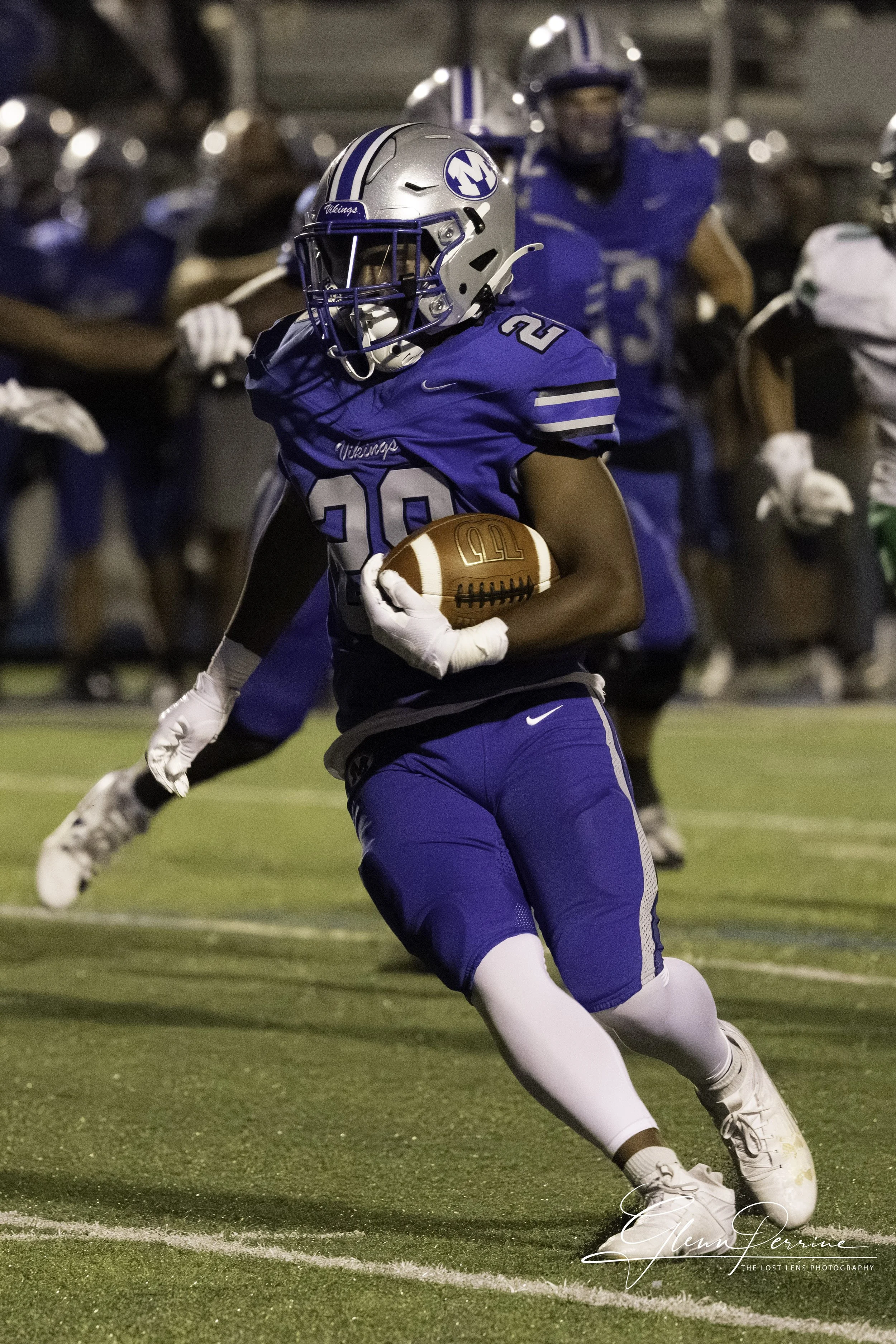 A football player in a blue uniform with the number 20 runs with the ball on the field during a game at night, with other players in the background.