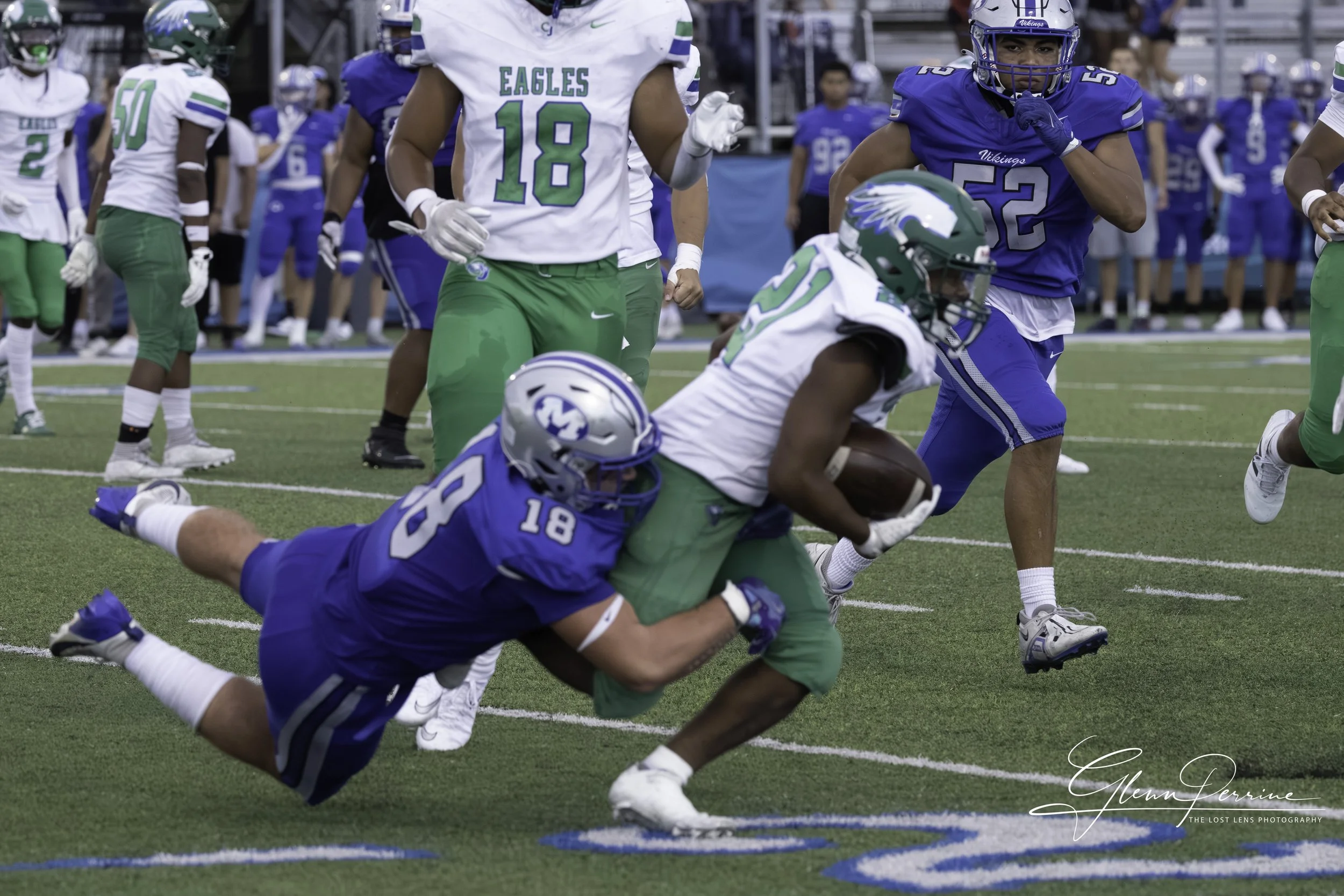 A football game with players in blue, green, and white uniforms. One player in a white and green uniform is running with the ball, tackled by a player in a blue uniform. Other players and spectators are visible in the background.