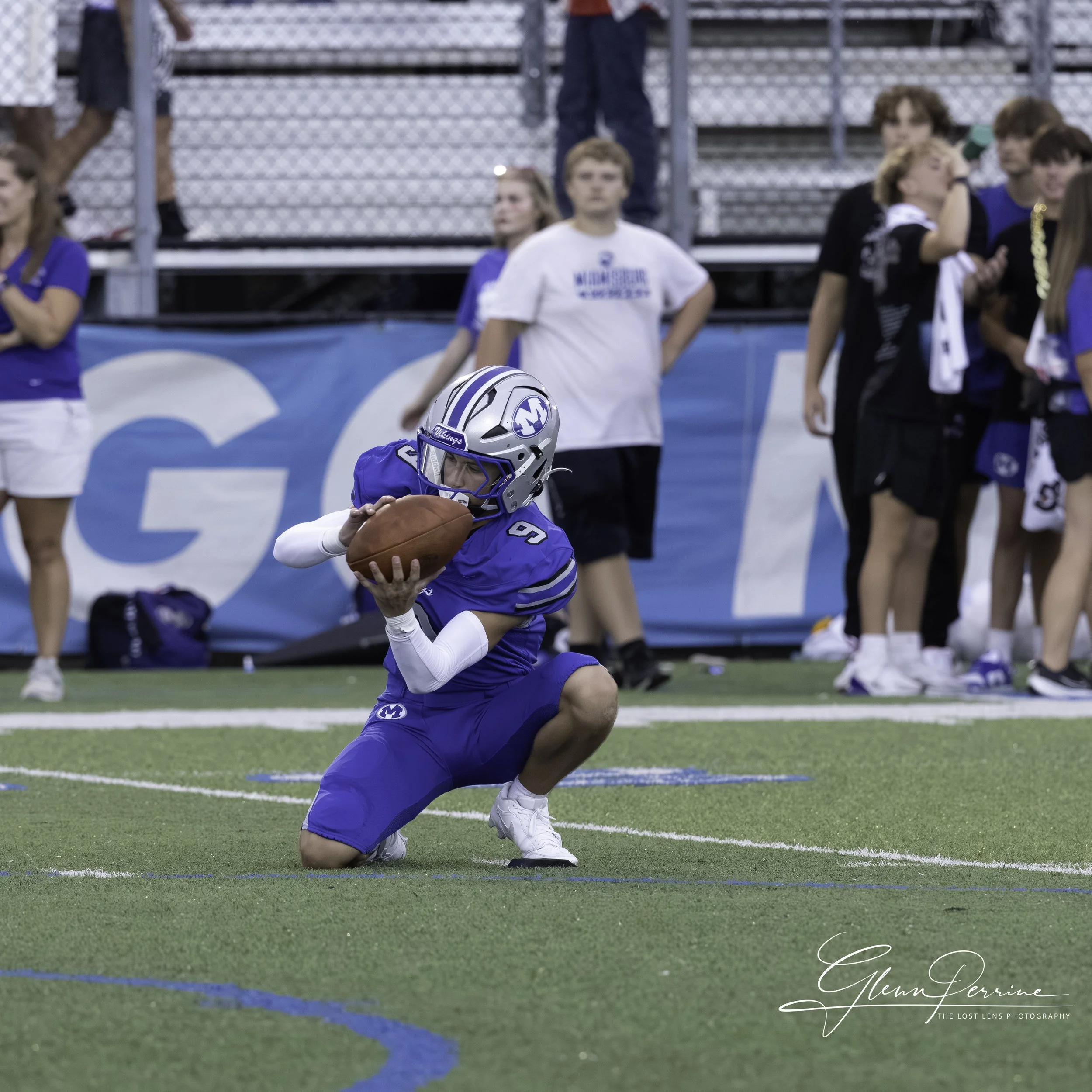 A football player in a blue uniform crouching on the field, holding a football, with people watching from the sideline in the background.