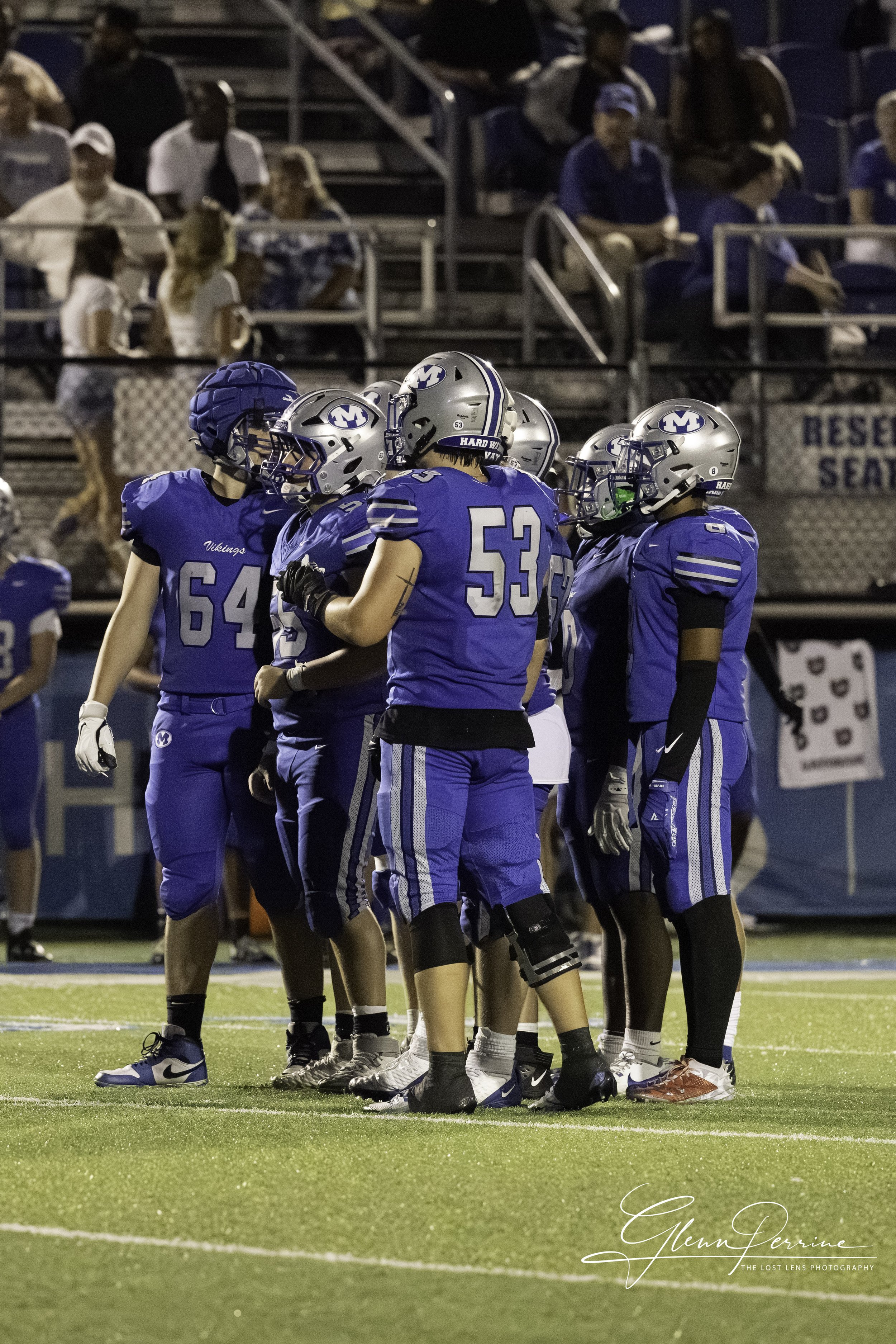 Football players in blue jerseys huddling on the field during a game, with spectators watching from the bleachers in the background.
