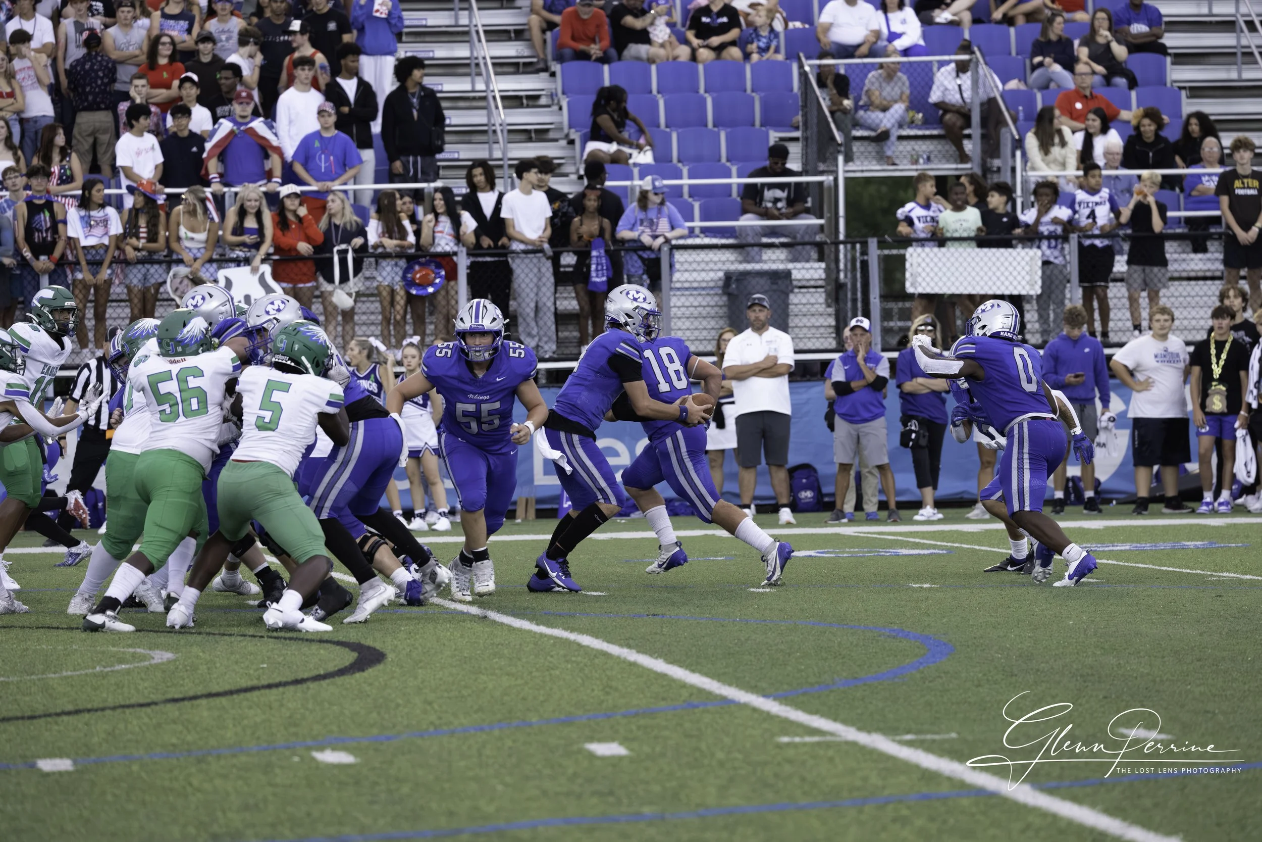 A football game underway with players in blue and green uniforms on the field, spectators watching from the bleachers in the background.