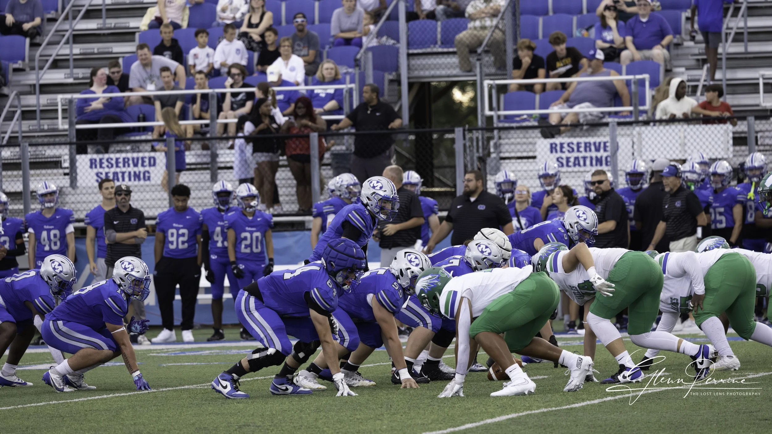 A football game with players on the line of scrimmage, wearing blue and green uniforms, on a green field with spectators in the background.