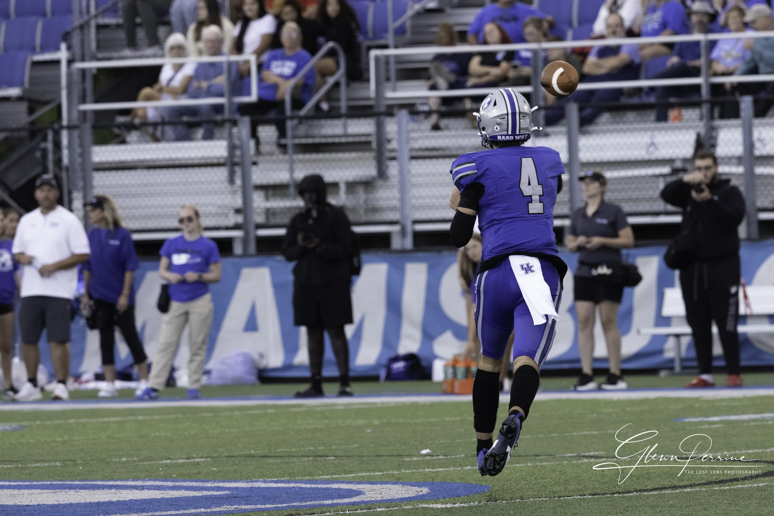 A football player in a blue uniform with the number 4 on his back, running on the field during a game. The player is wearing a helmet and black arm sleeves. Spectators are watching from the stands in the background.