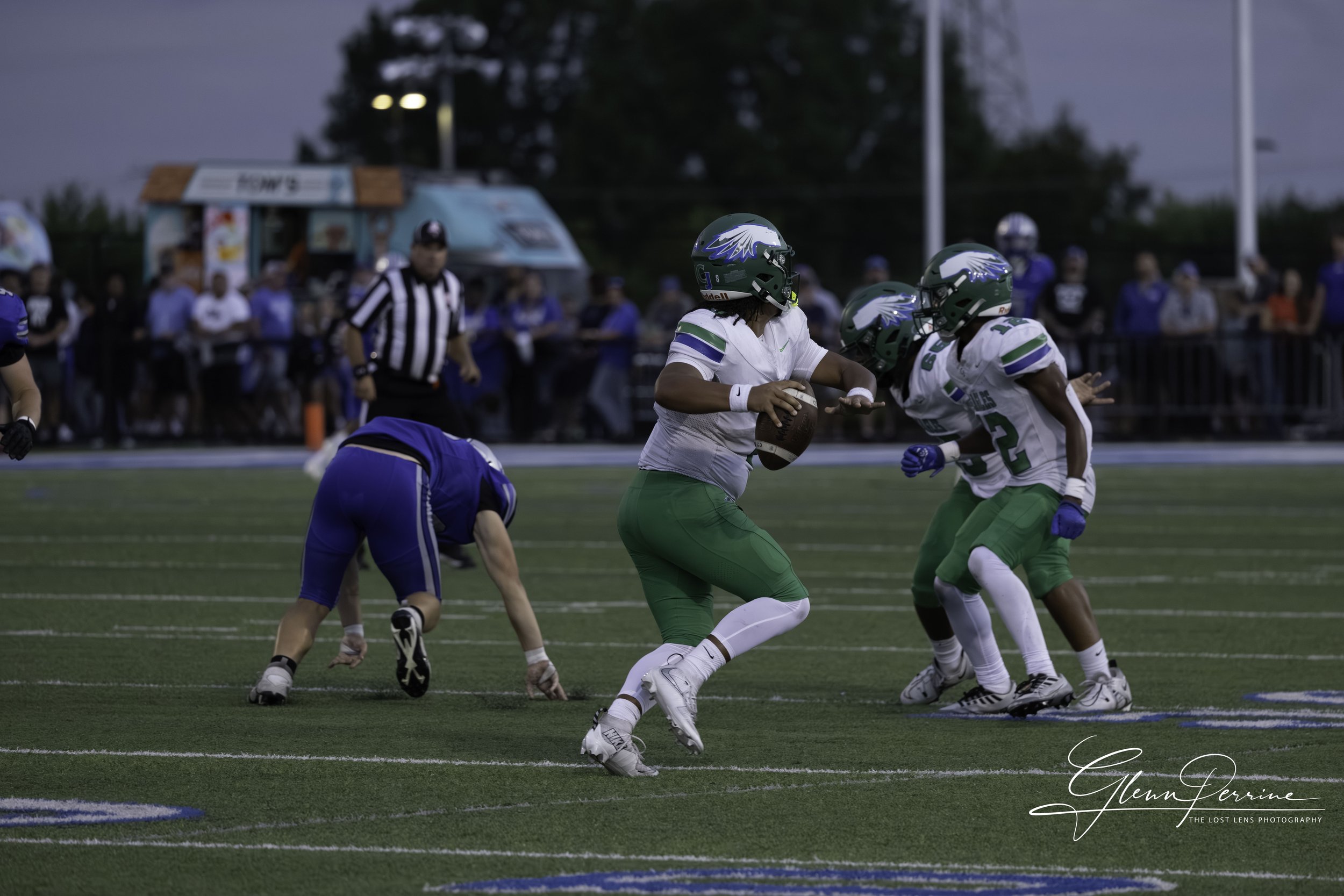 American football players in white and green uniforms on the field, with one player holding the ball, preparing to run. Opposing team player in blue is bent over on the ground, with a crowd of spectators and a food truck in the background.