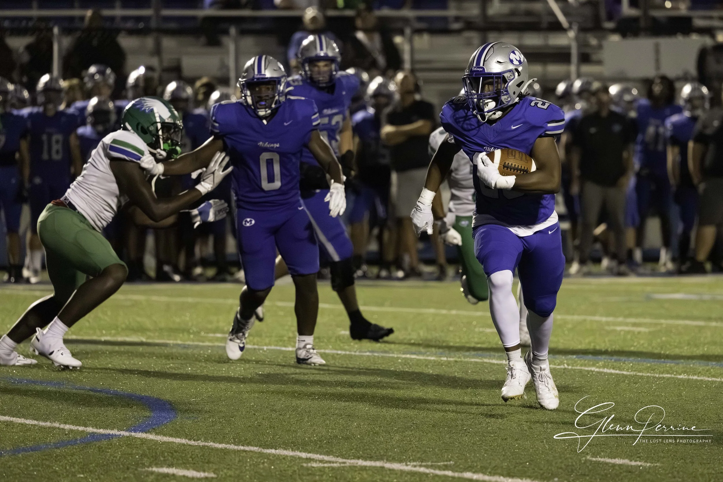 High school football game with a player in blue running with the ball while being tackled by an opponent in green and white. Other players and spectators are in the background.