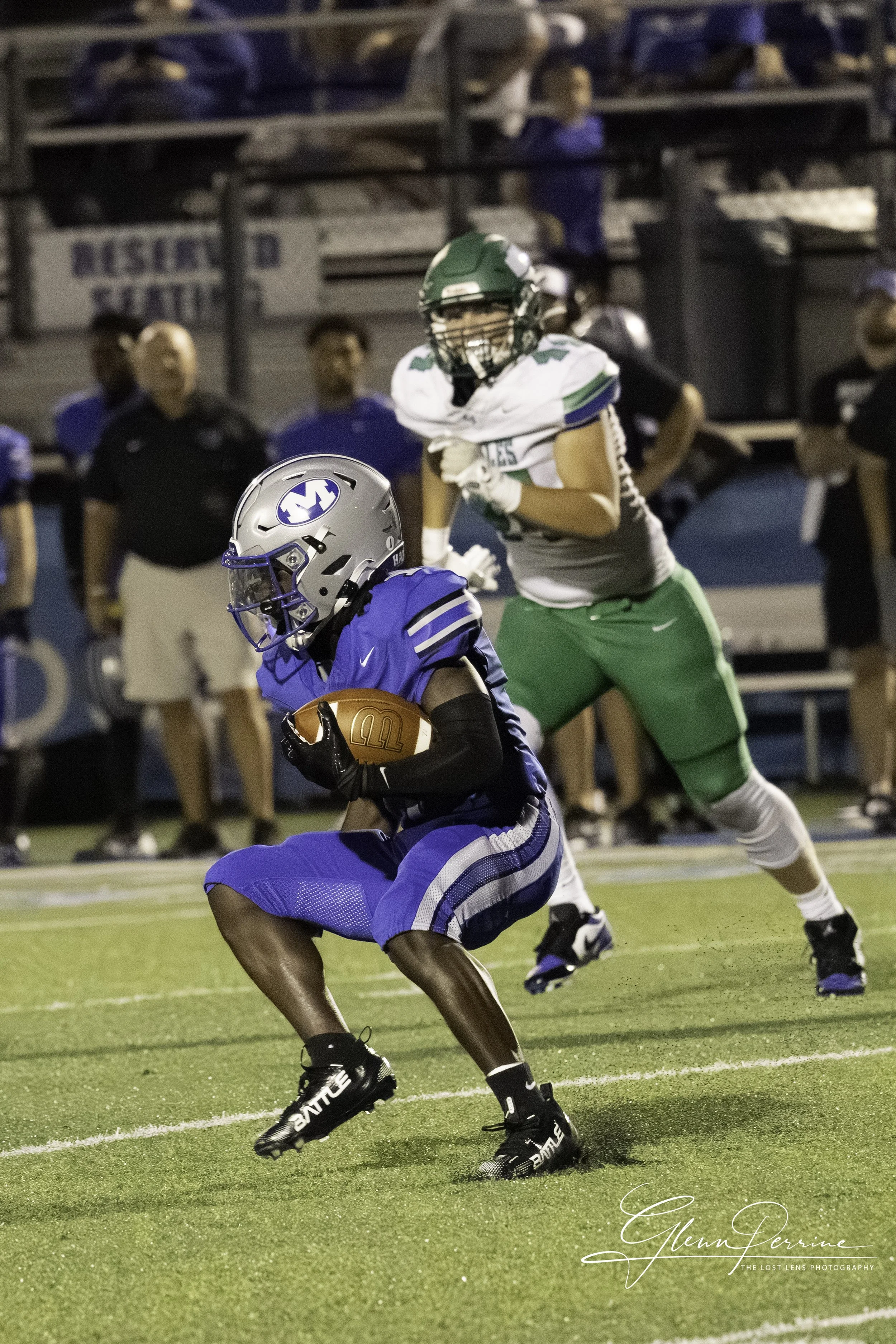 A football player in a blue uniform holding the ball runs on a field while a player in a white and green uniform chases him, with spectators and coaches in the background.