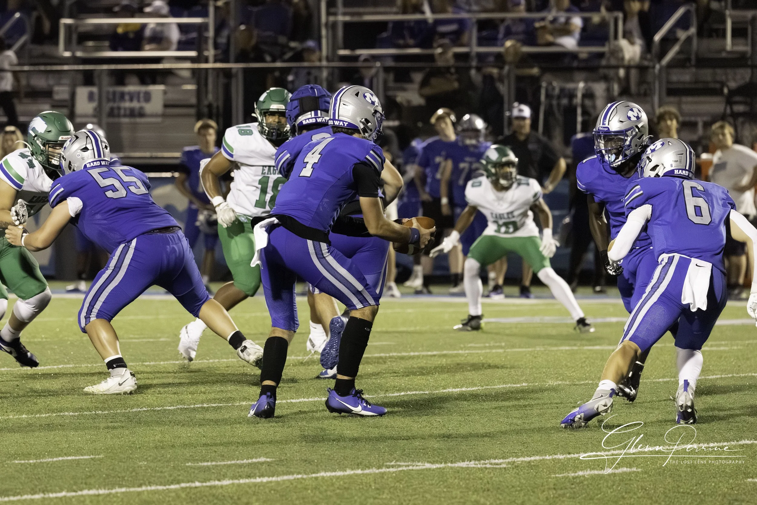 A high school football game with players in blue and green uniforms, some running with the ball and others blocking, on a well-lit field during evening.