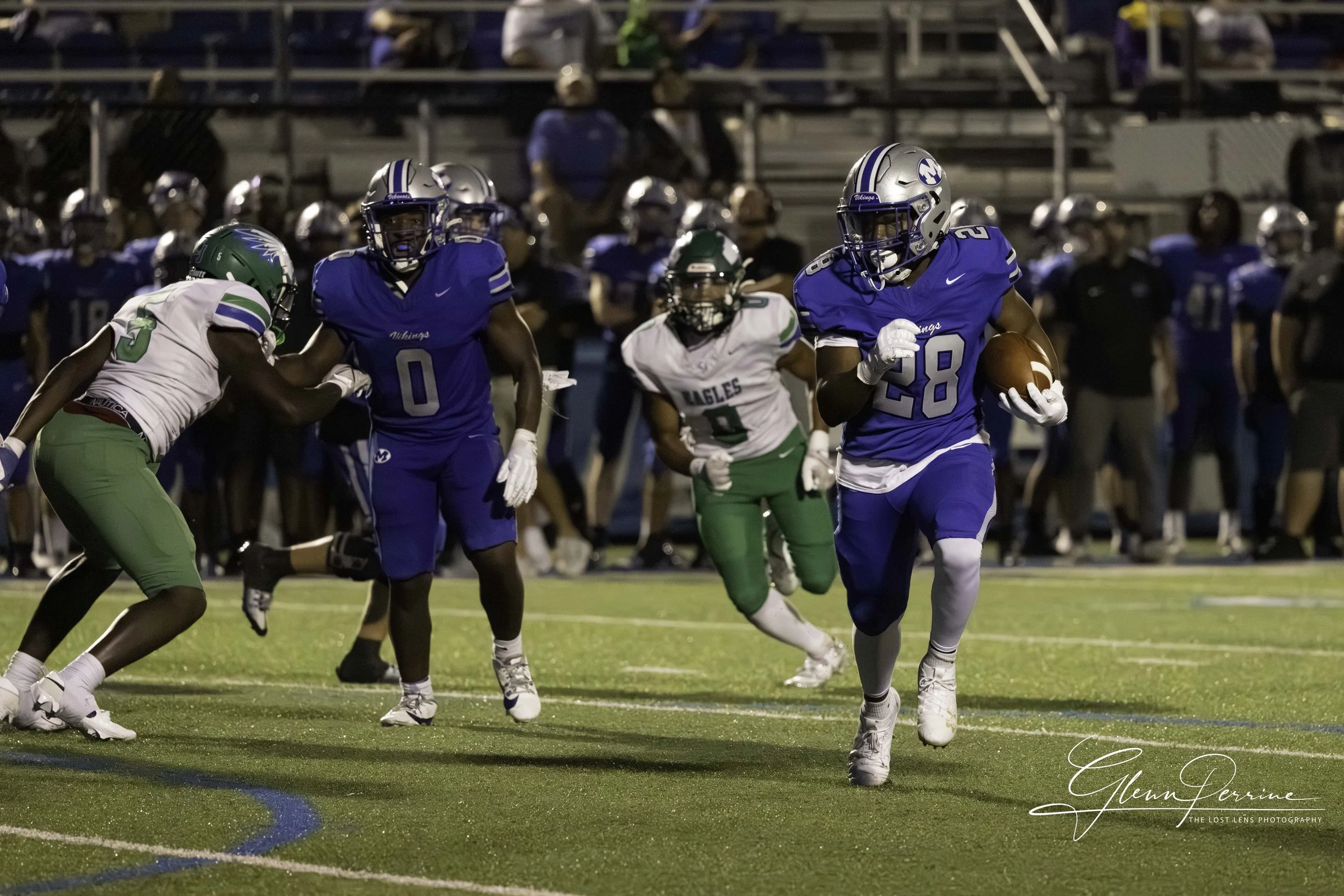 A football game with players in blue and green uniforms. A player in a blue uniform holding a football runs while being pursued by a player in a green uniform. Other players and spectators are visible in the background.