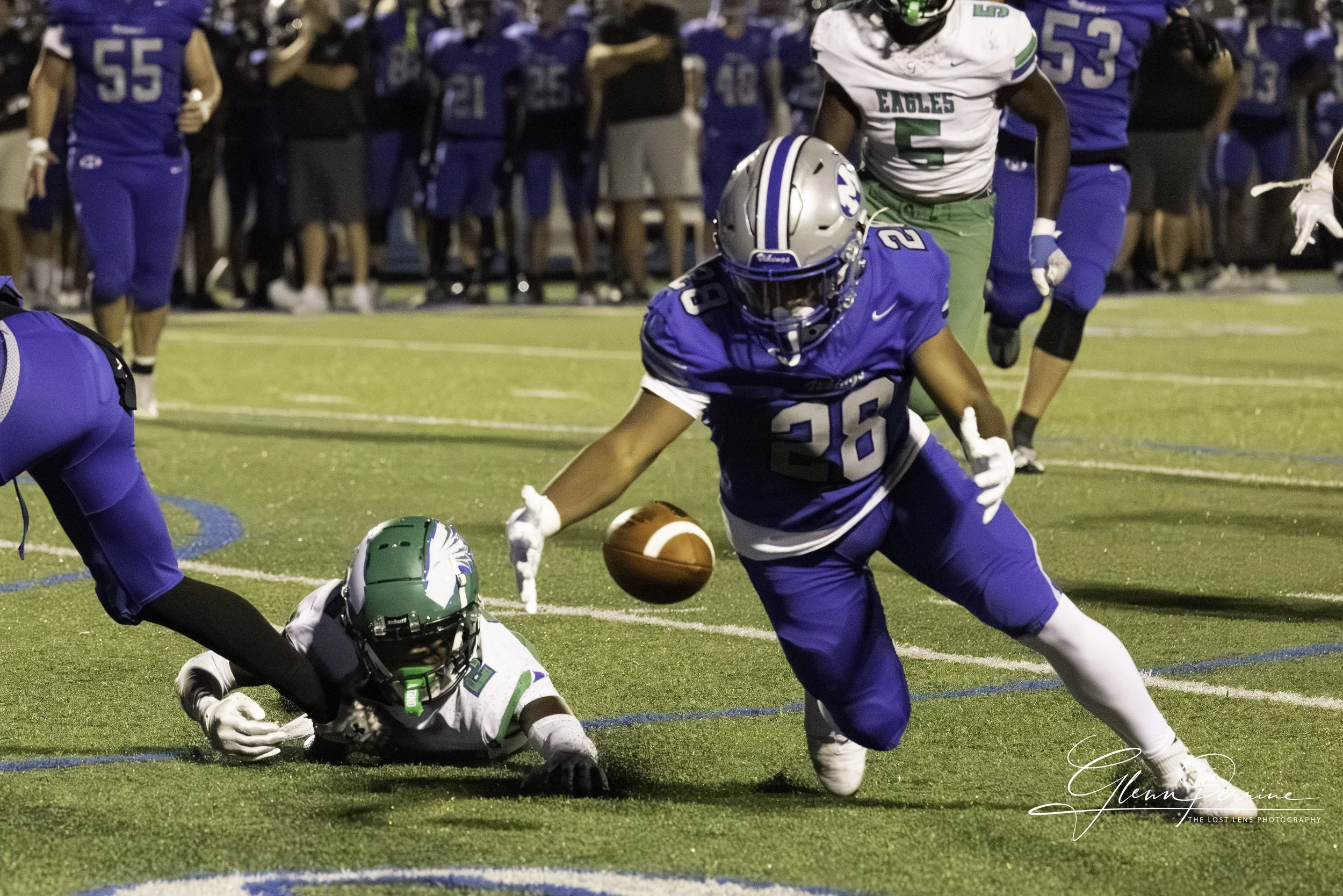 A football game scene where a player in a blue jersey with the number 26 is diving to recover a fumbled football on the field, with a player in a white and green jersey lying on the ground nearby. Other players and spectators are visible in the backg