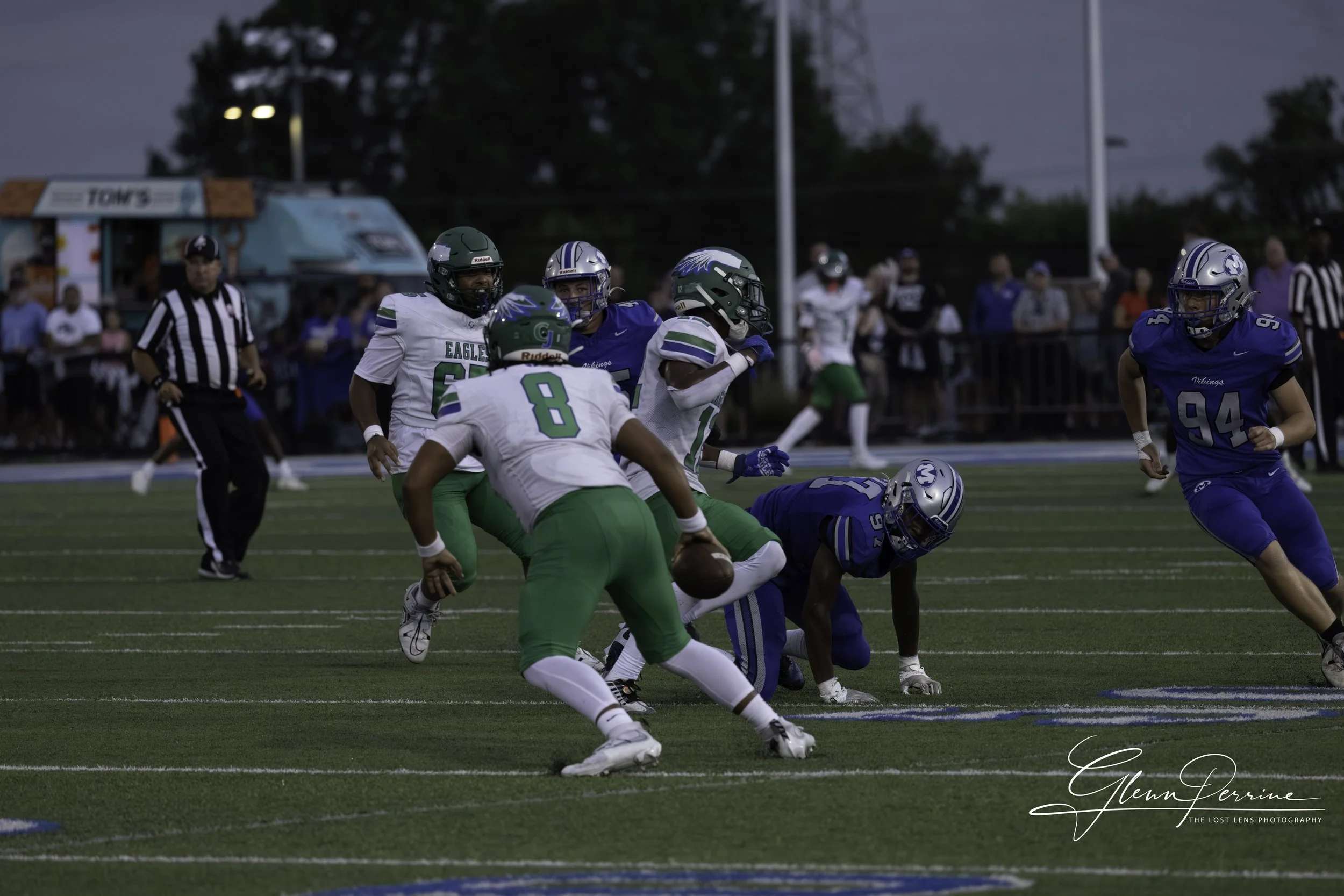 A football game with players in green and white uniforms and players in blue uniforms. A player in white and green is running with the ball while others try to block or tackle. Spectators watch from the sidelines.