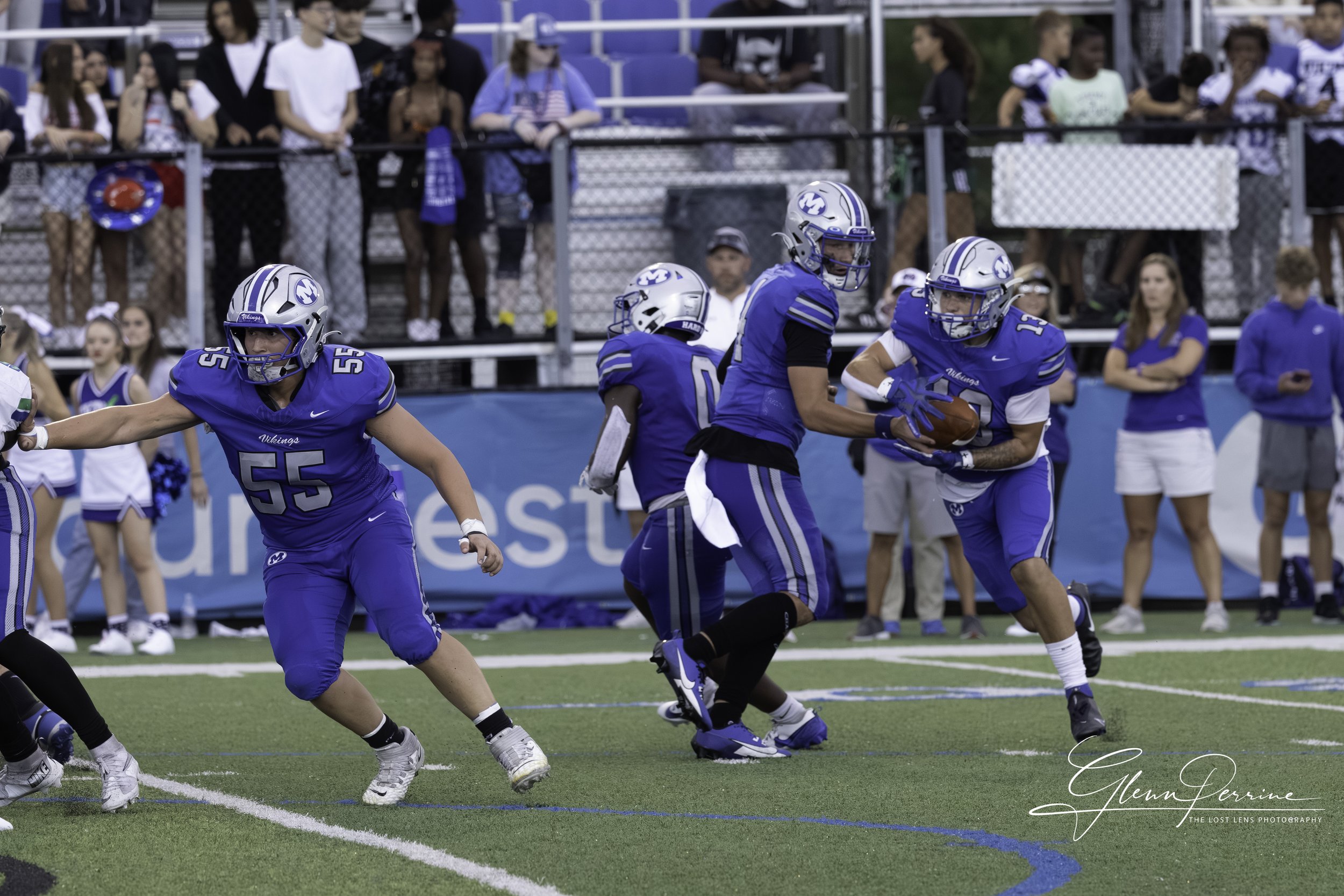 A football game showing players in blue uniforms with whites, with some holding the football, during a game on a field with spectators in the background.