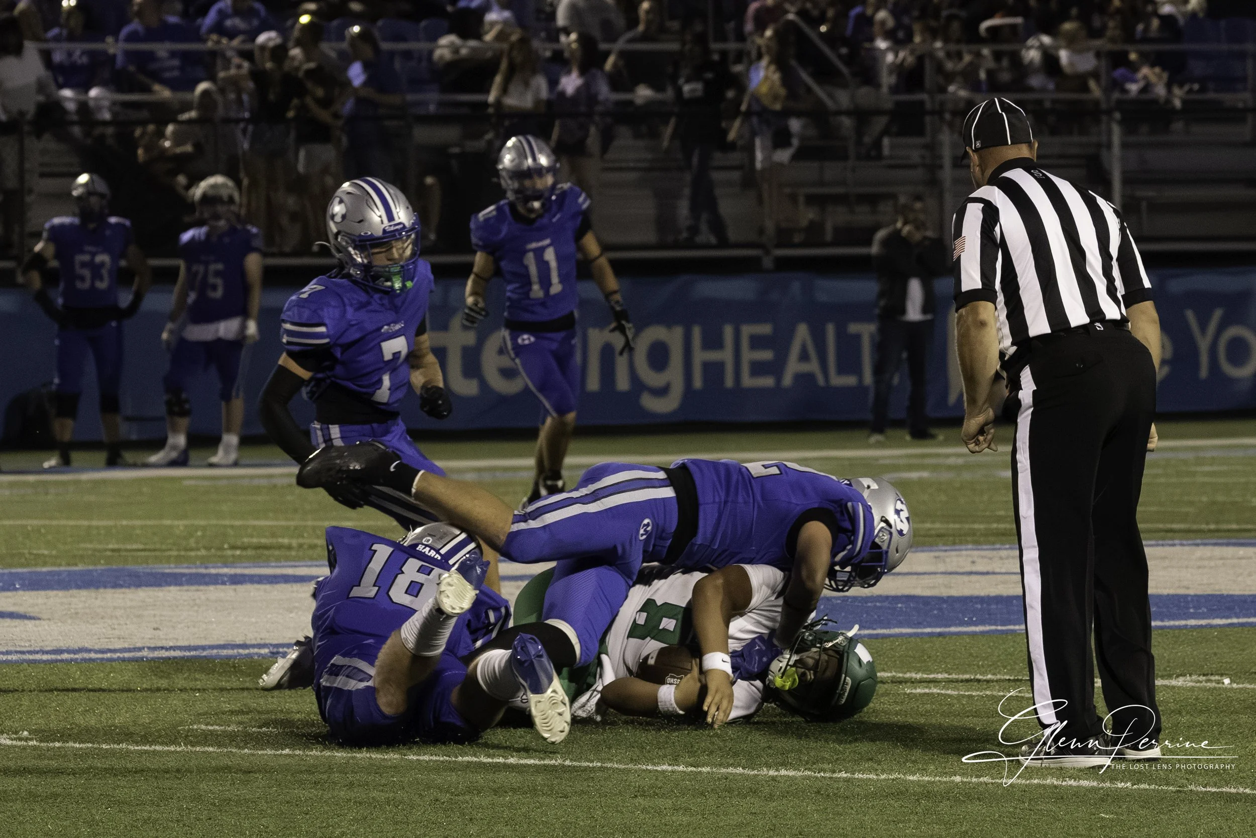 Football players in blue uniforms tackle a player in a green and white uniform on the field during a game. An official in a black-and-white striped shirt watches nearby. Spectators are in the background.