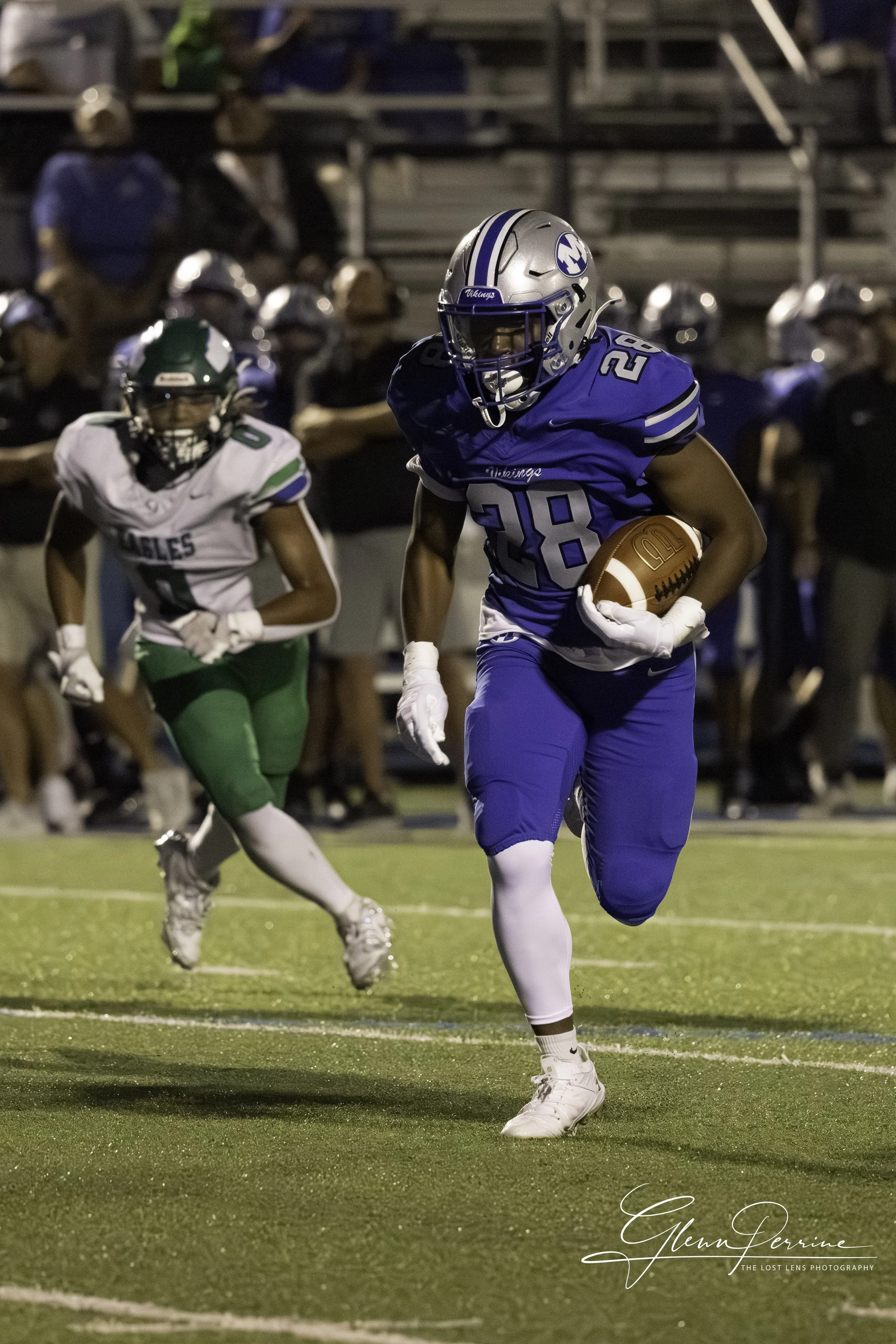 A football player in a blue uniform with the number 28 runs with the ball, pursued by a player in a green and white uniform during a game at night.