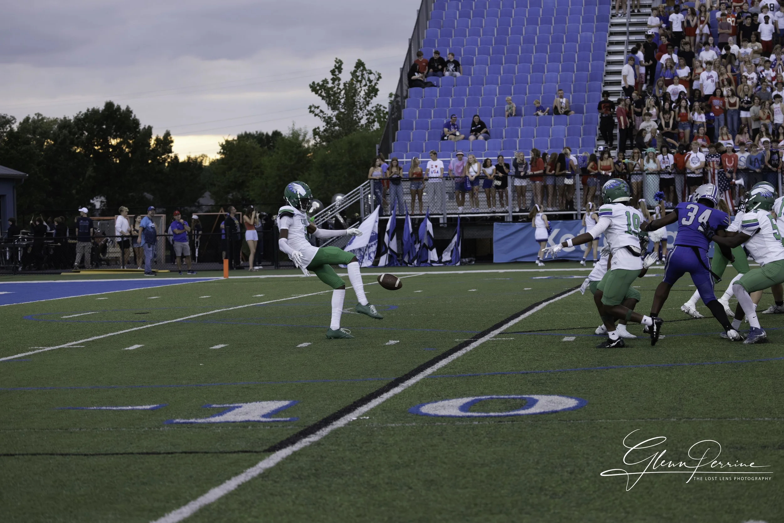 A football player kicks a football on the field as teammates block opponents, with spectators seated in the stands watching the game.