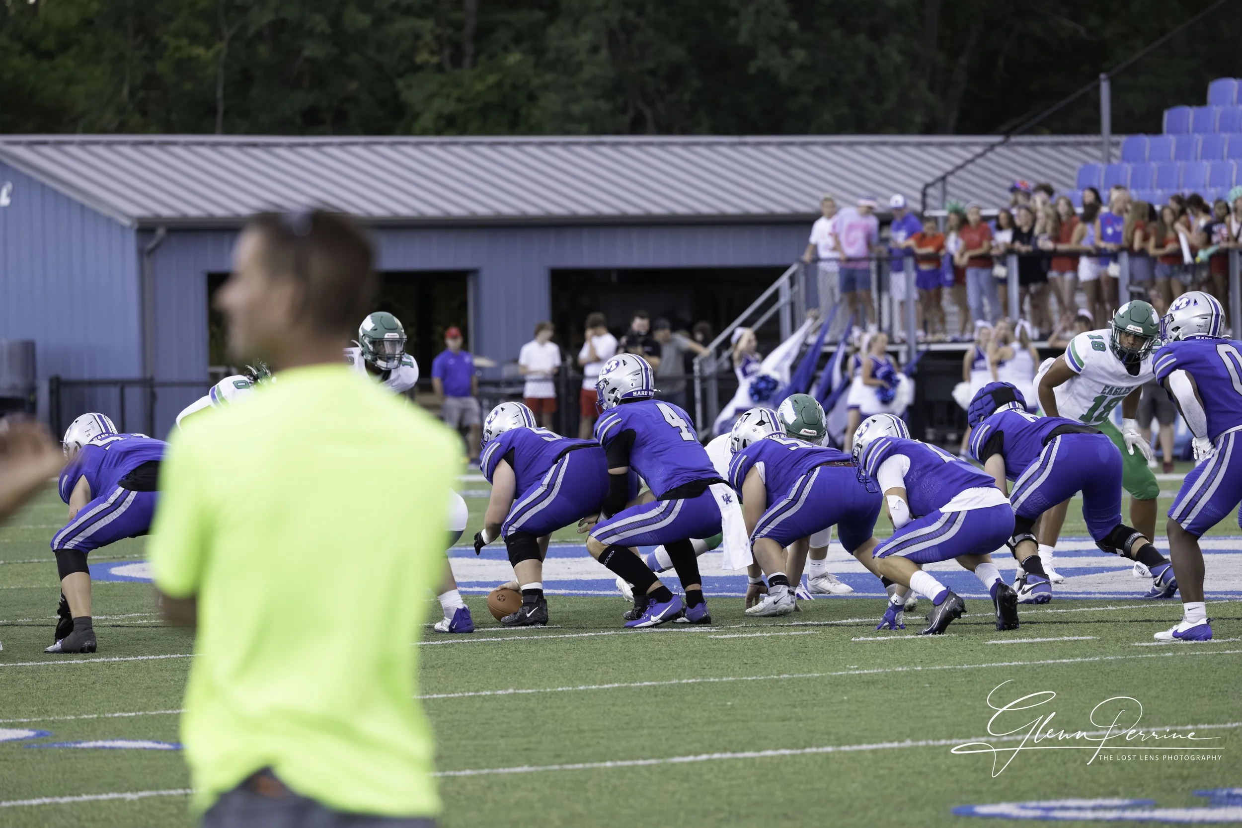 Football players in blue and white uniforms line up at the line of scrimmage on a football field during a game, with spectators watching from the stands.
