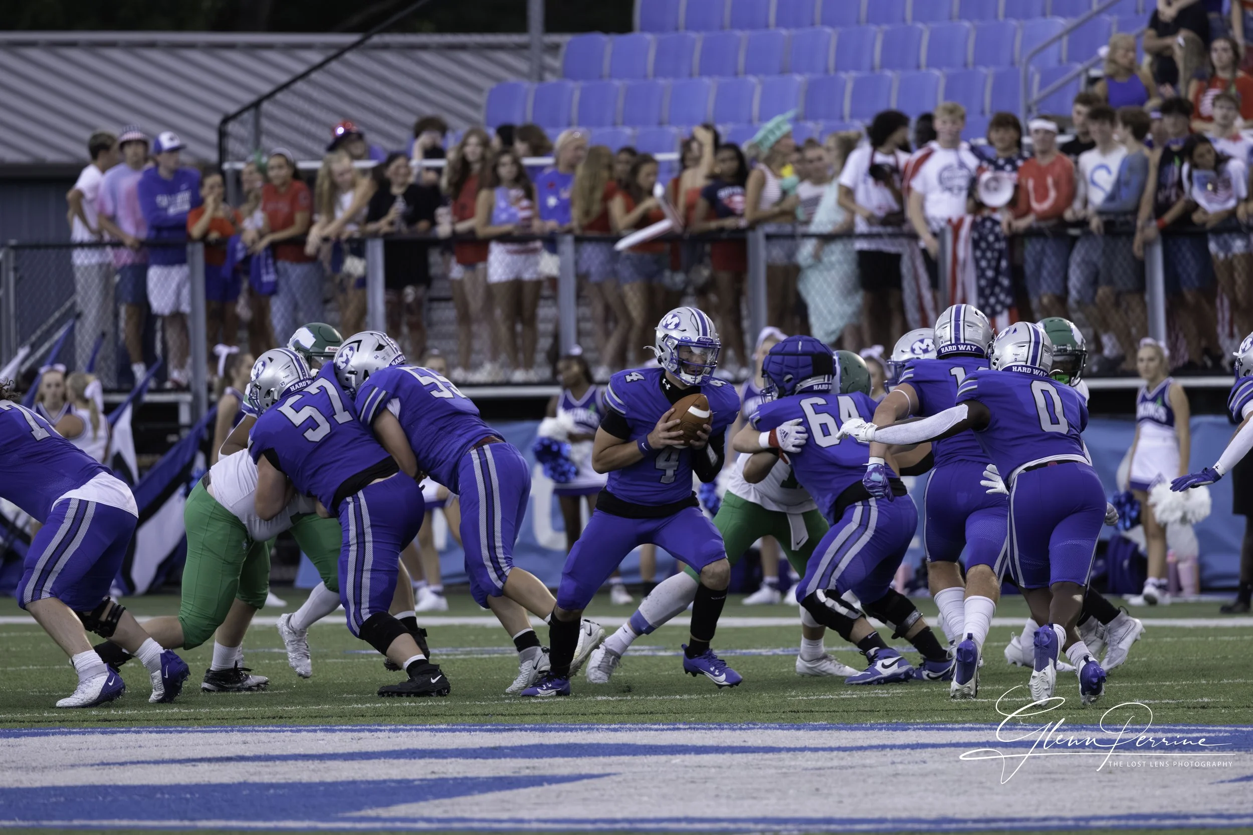 A youth football game with players in blue and white uniforms on the field, with the quarterback holding the football and other players blocking. Spectators are standing behind the fence in the background.