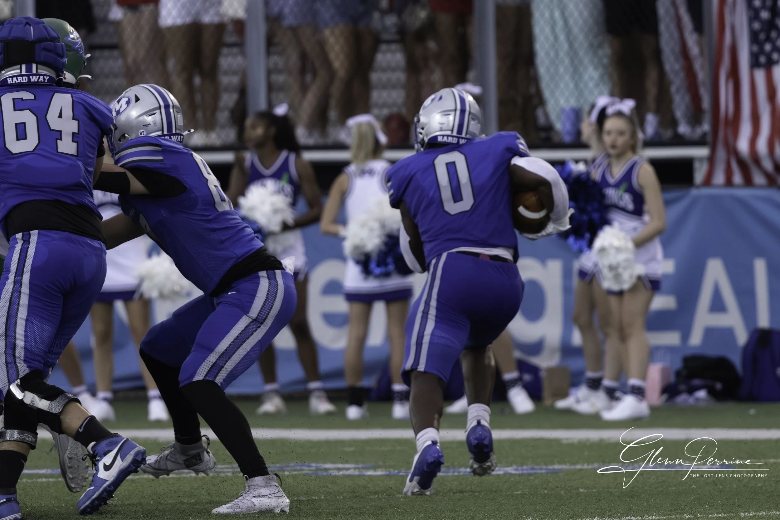 Young football players in blue uniforms and helmets playing a game, with cheerleaders and spectators in the background.