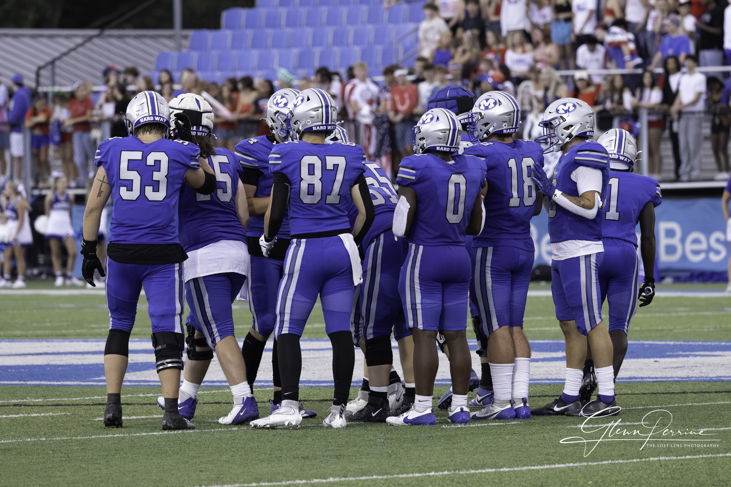 A group of American football players in blue uniforms huddling on the field during a game, with spectators watching from the stands.