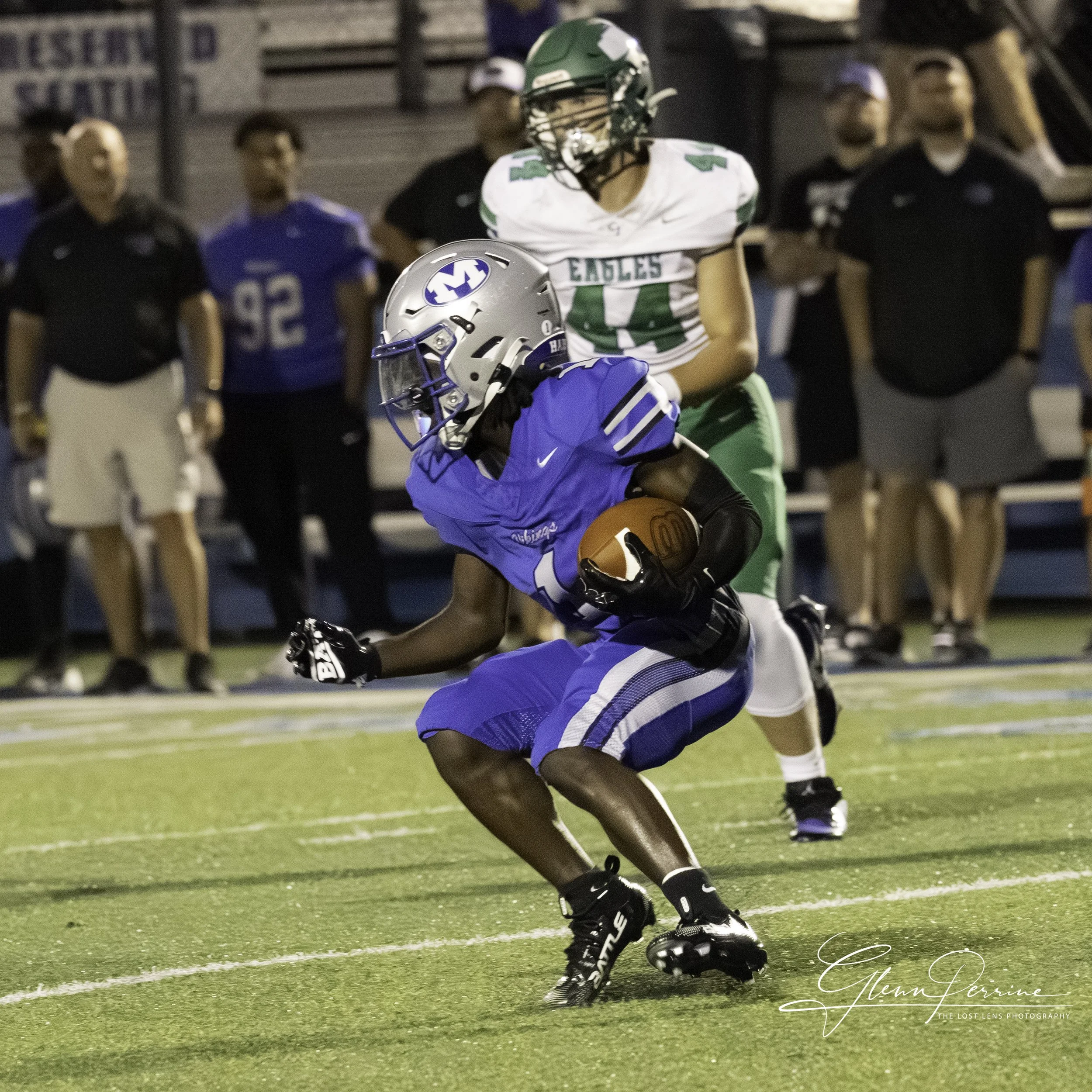 A football player in a blue uniform and helmet is running with the ball on the field, with a player in a white and green uniform chasing behind him. Several spectators and team staff are visible in the background.