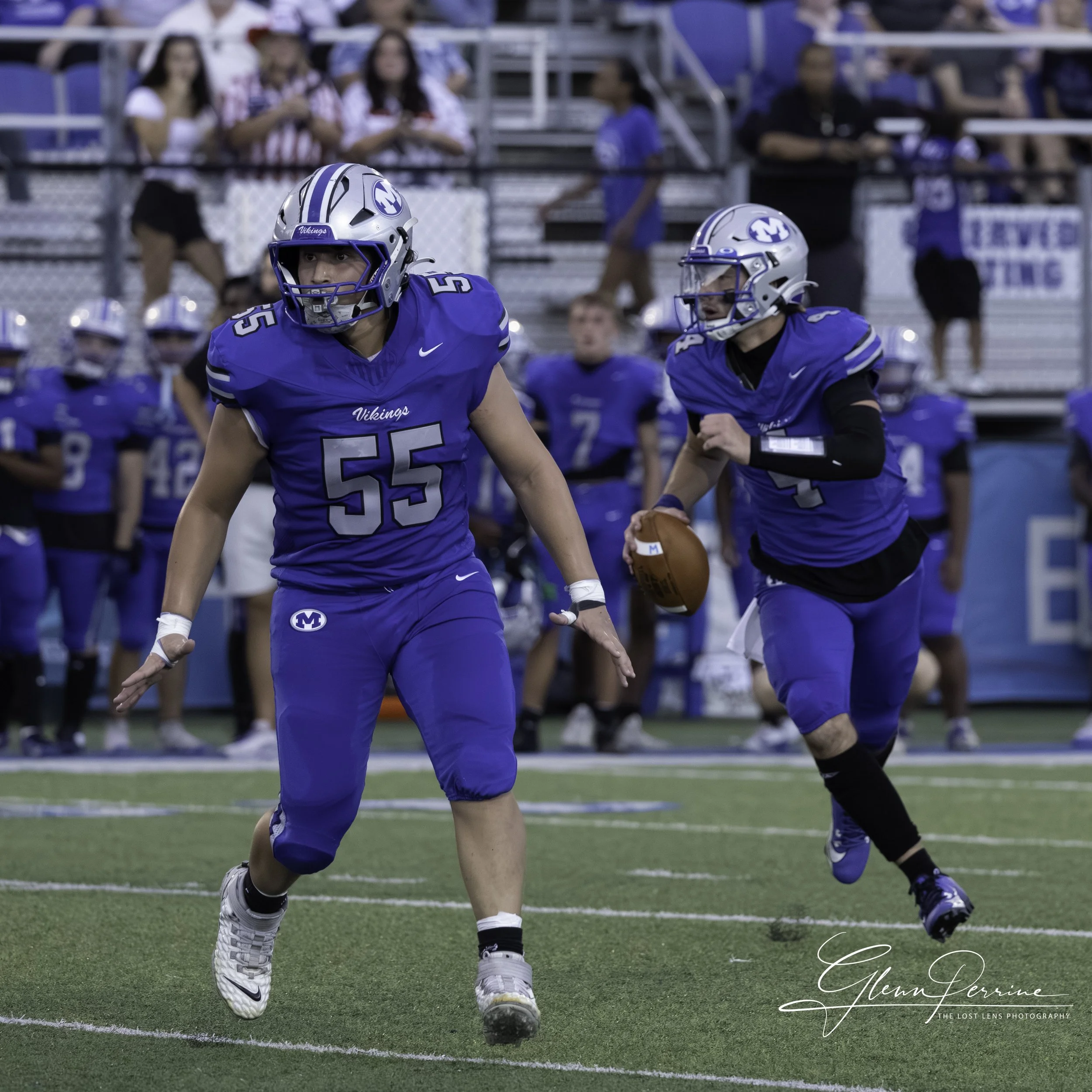 Football players in blue uniforms on field, one running with ball, team bench and spectators in background.