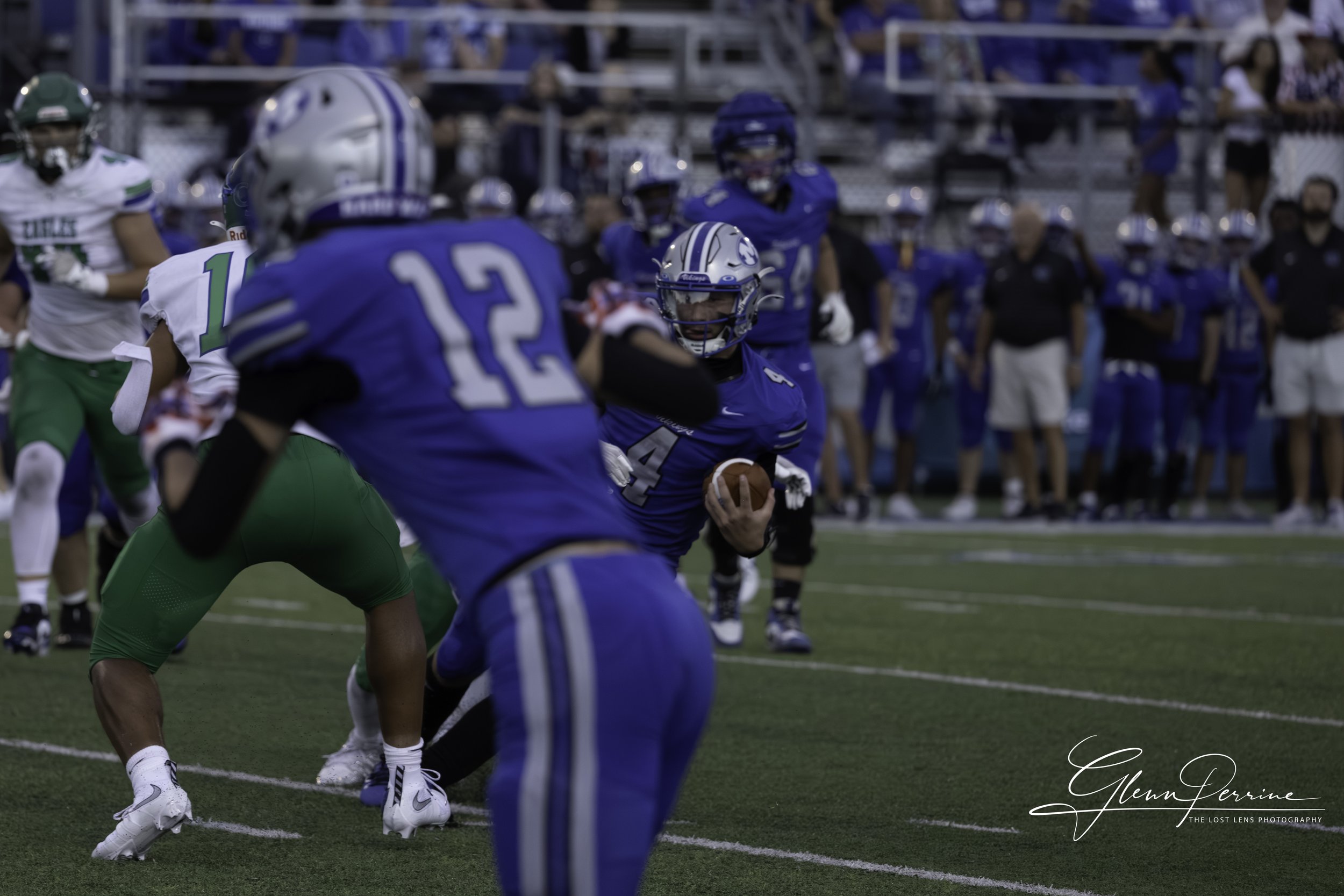 A football game with players in blue and green uniforms on the field. The player in blue holding the football is preparing to run, while other players are in position or engaging. Spectators are visible in the background.