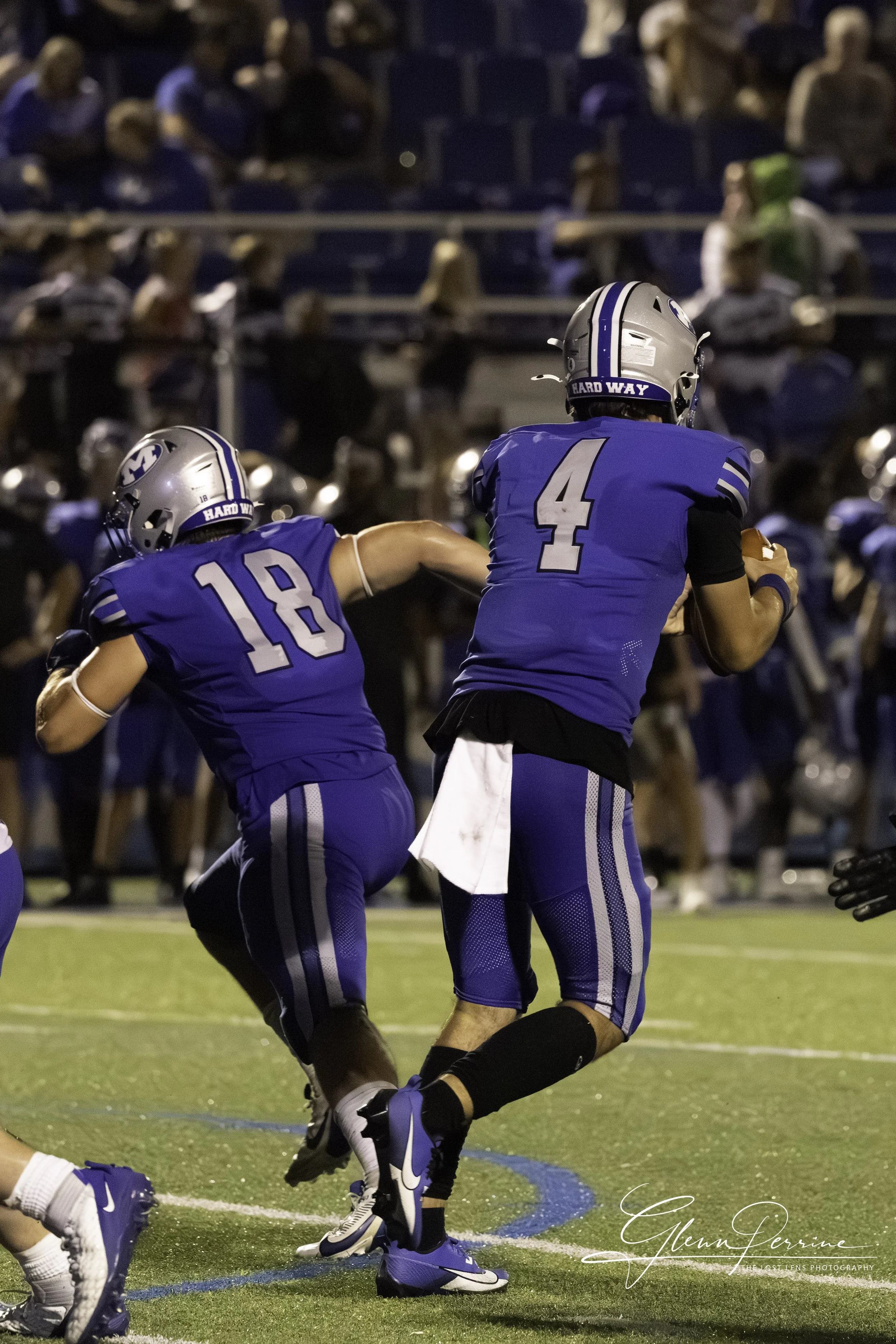 Two football players in blue uniforms and helmets on the field, with the player wearing jersey number 4 holding a football and running while the other player, number 18, appears to be blocking or supporting him. Spectators are seated in the backgroun