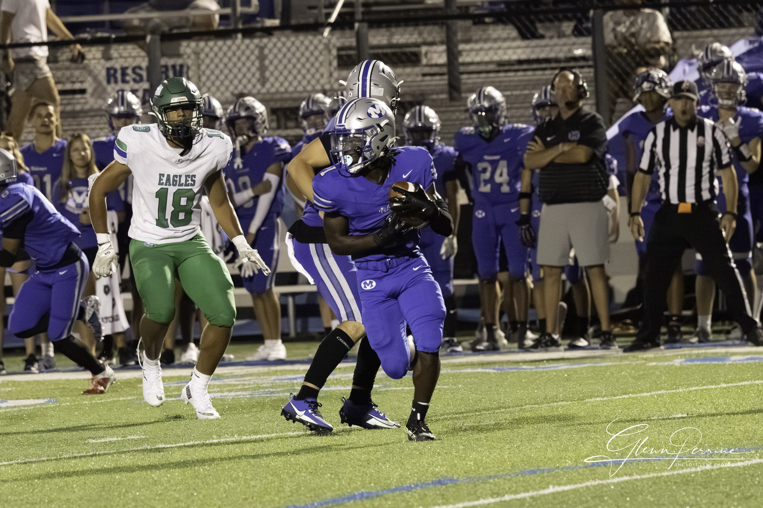 An American football game with players on the field, including one in a blue uniform holding the ball and running, and another in a white and green uniform chasing. Spectators and coaches are visible on the sidelines.