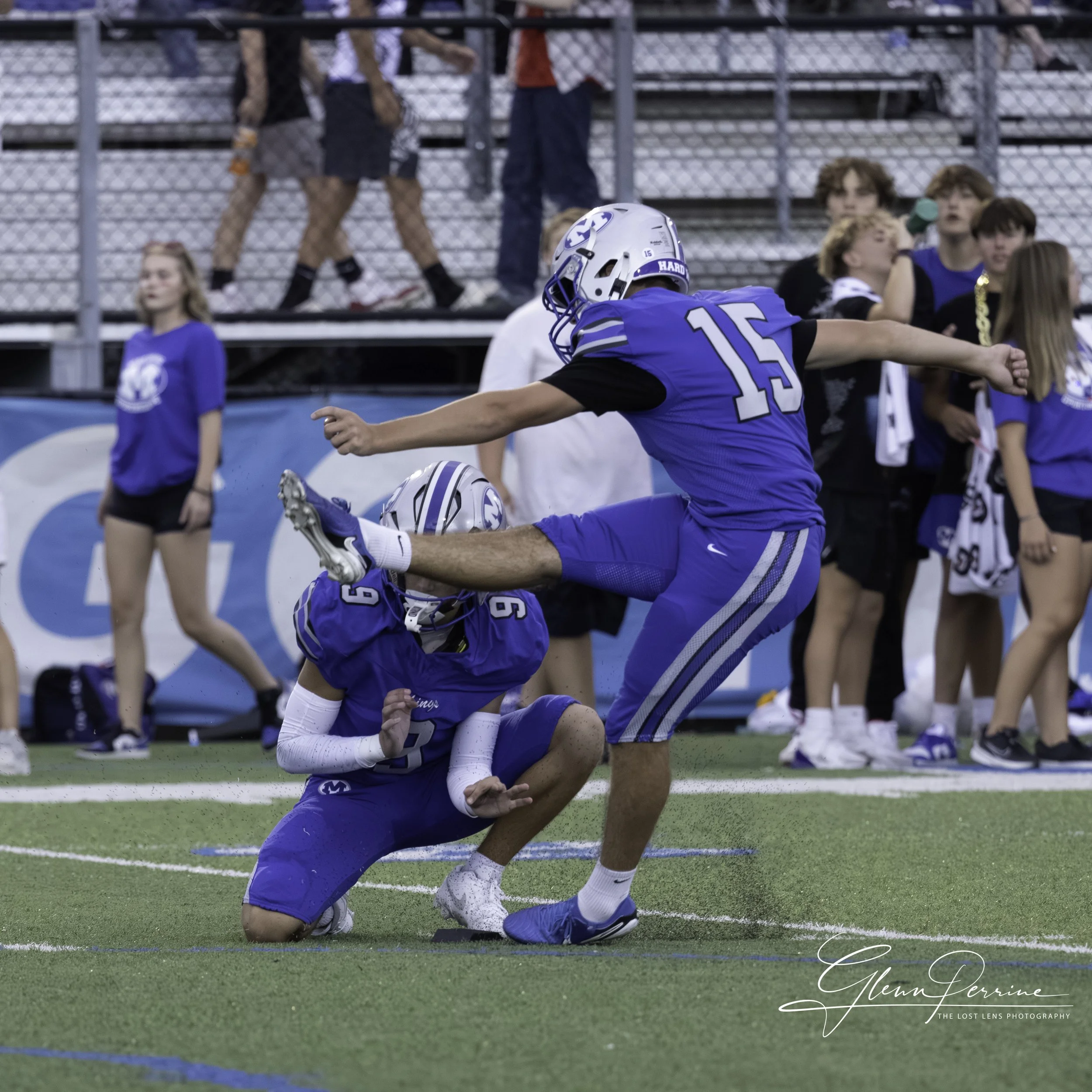 Two football players in blue jerseys celebrating on the field, one kneeling and the other kicking the other's helmet, with a group of spectators in the background.