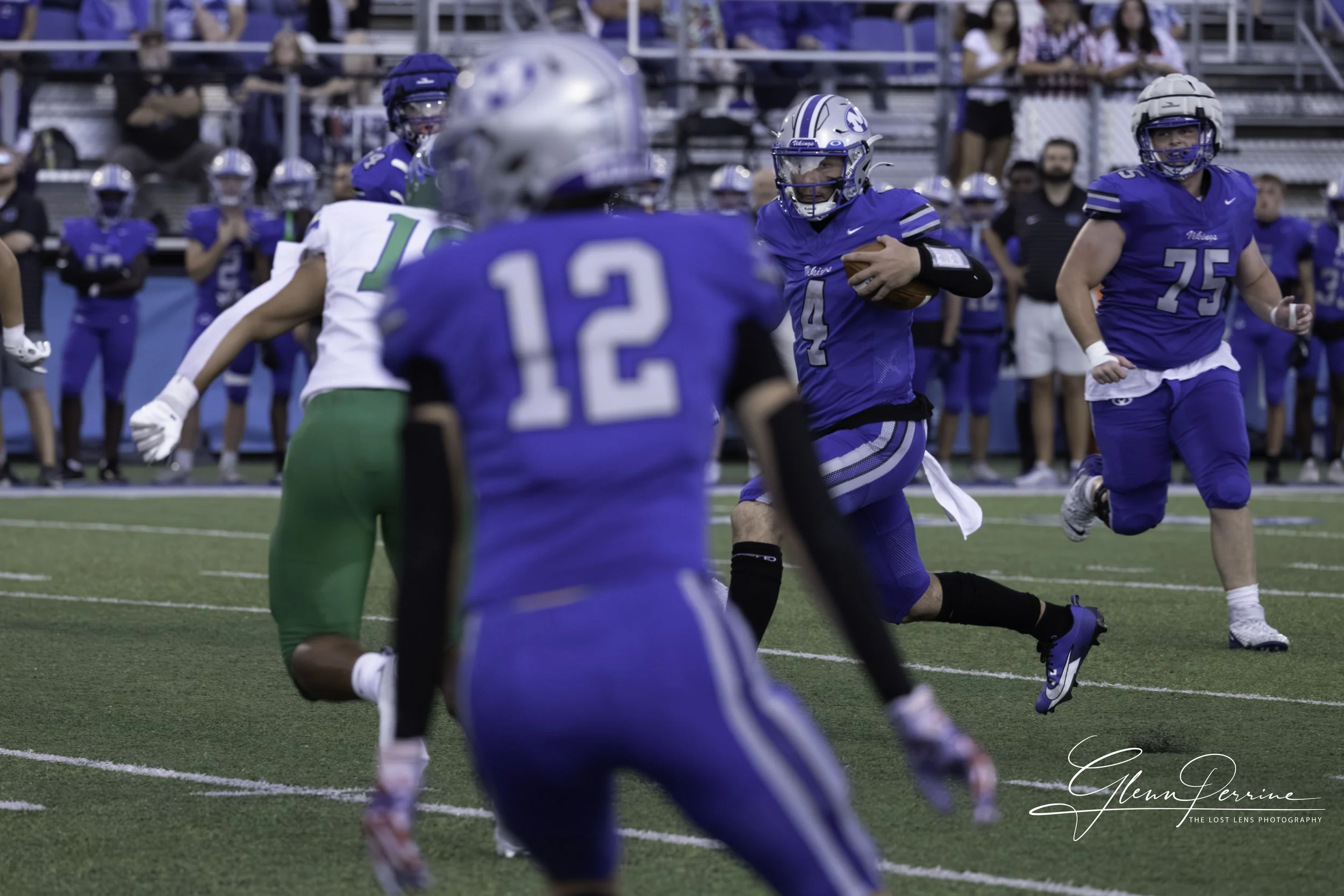 A football player in a blue uniform number 14 is running with the ball during a game. Several other players in blue uniforms and one in a white and green uniform are on the field, with spectators watching from the bleachers.