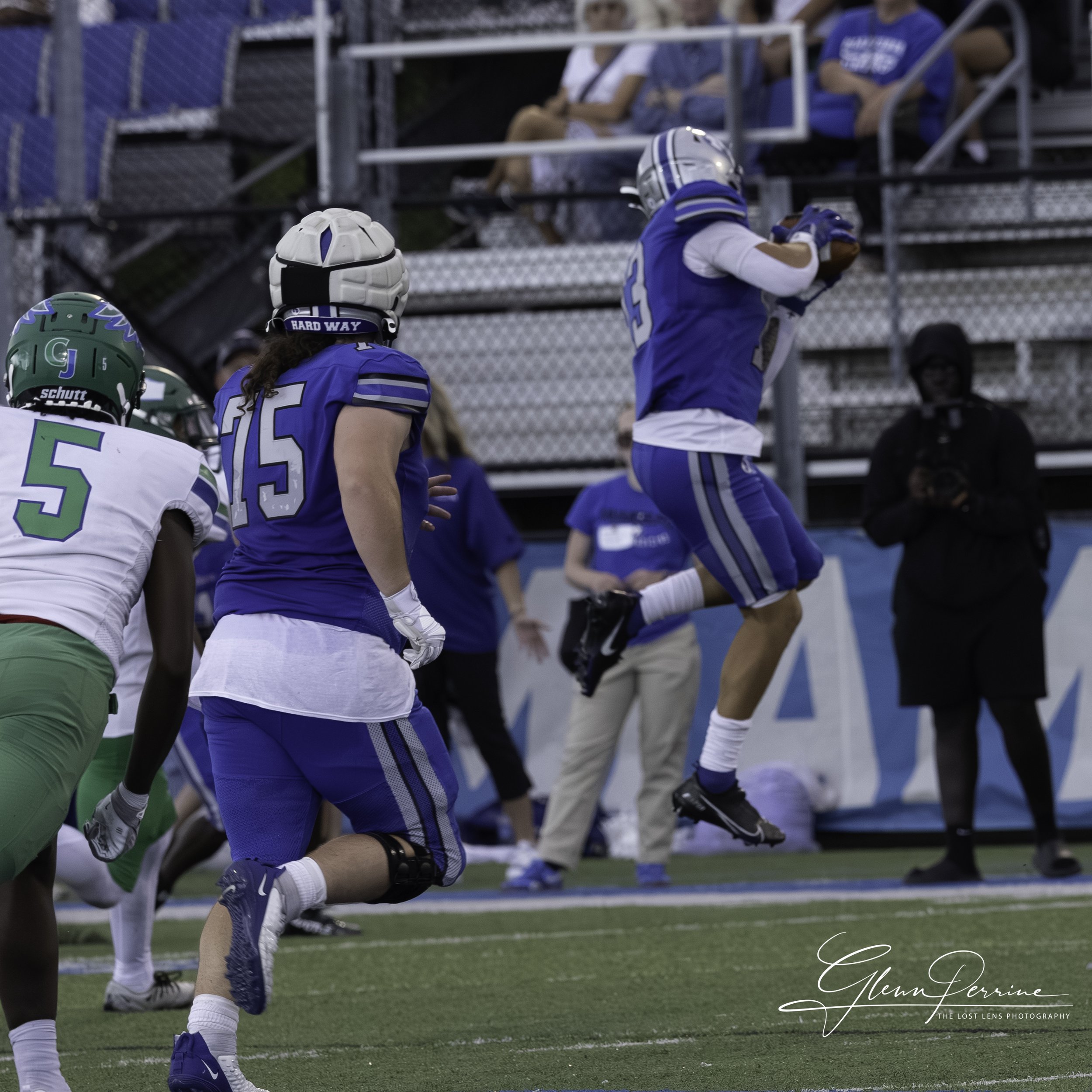 American football players during a game, with one player in blue jumping to catch the ball while others run on the field, and spectators in the stands watching.