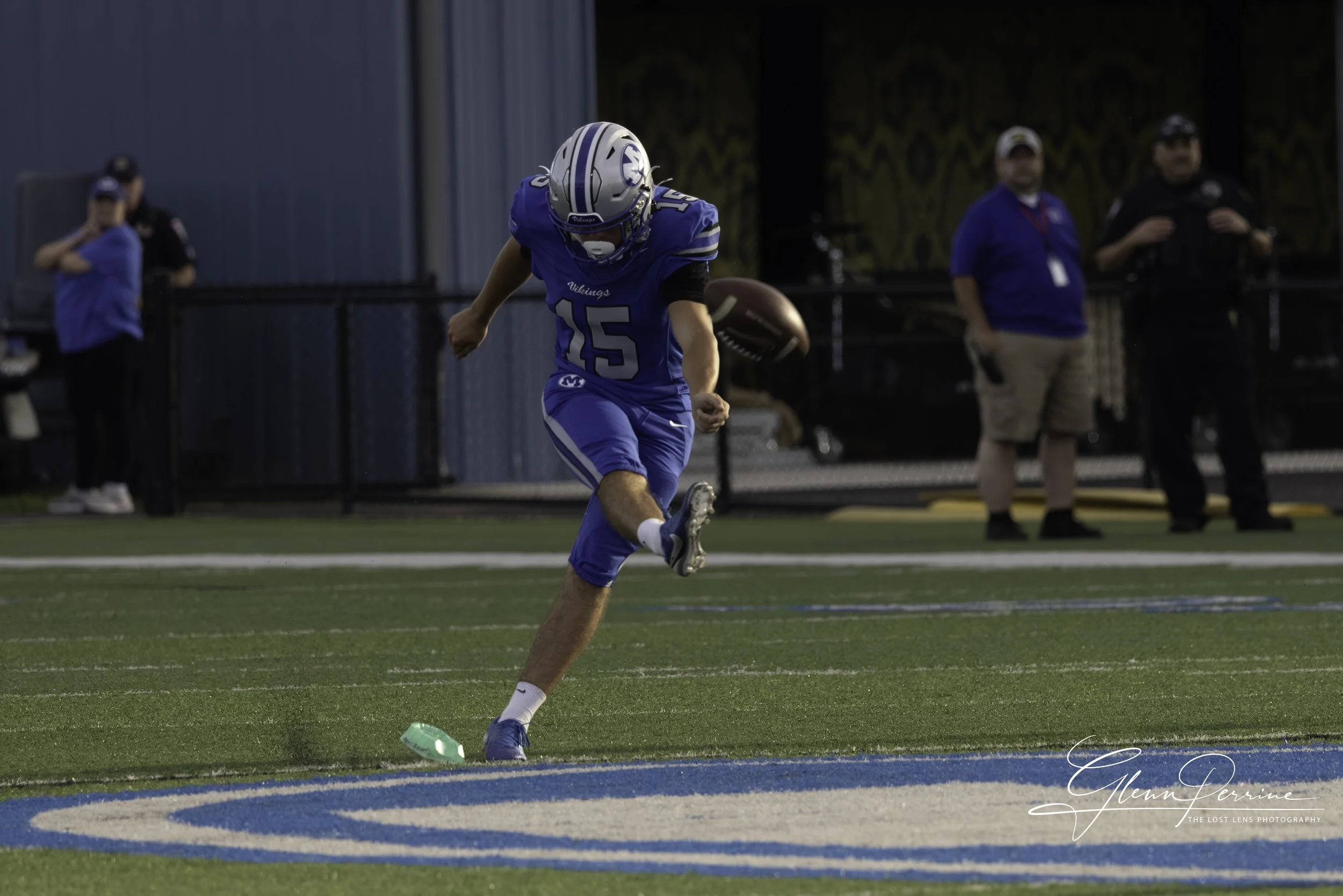 A football player in a blue uniform with the number 15 is on a field, kicking the football. There are three people in the background, two wearing blue shirts and one in black attire. The field has green turf and a decorated background.