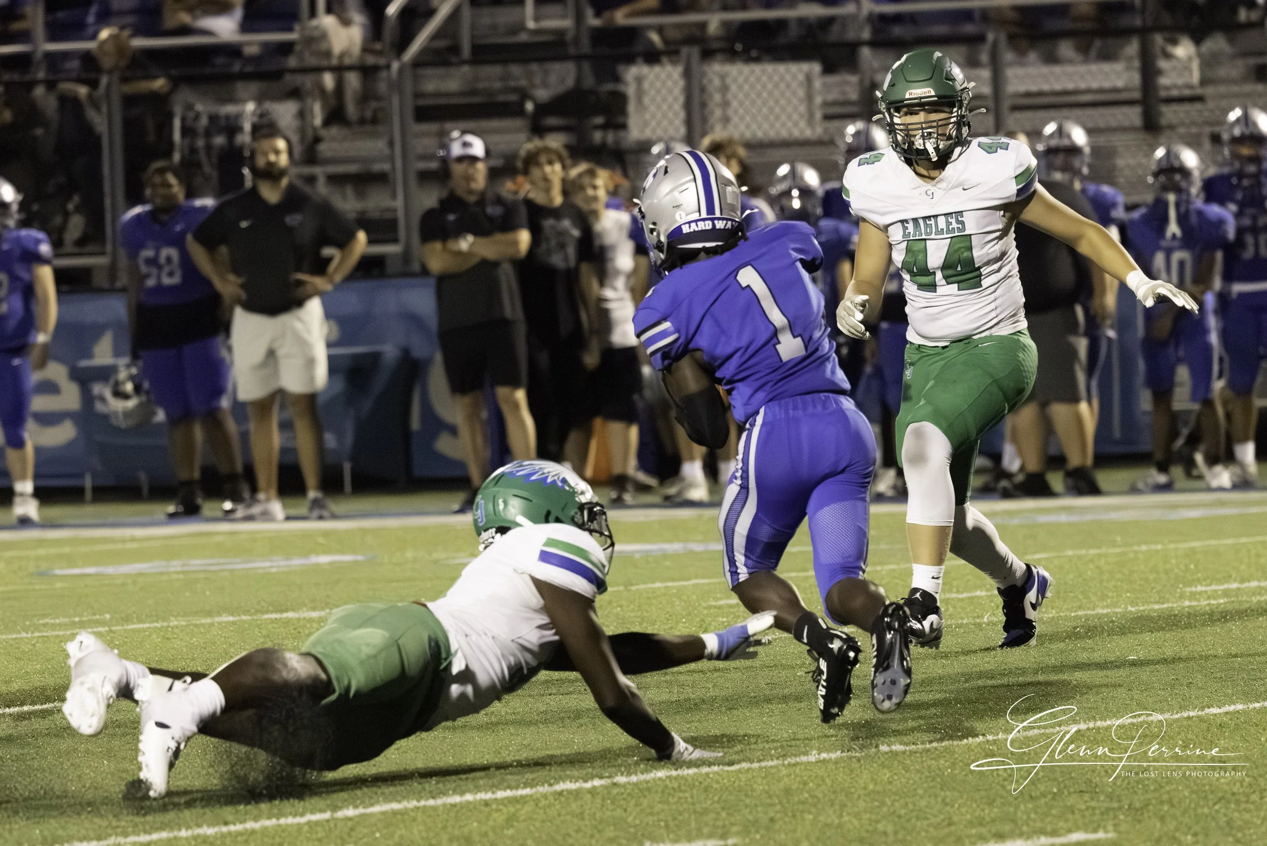An American football game with a player in a blue uniform running while being tackled by a player in a white and green uniform, with other players and spectators in the background.