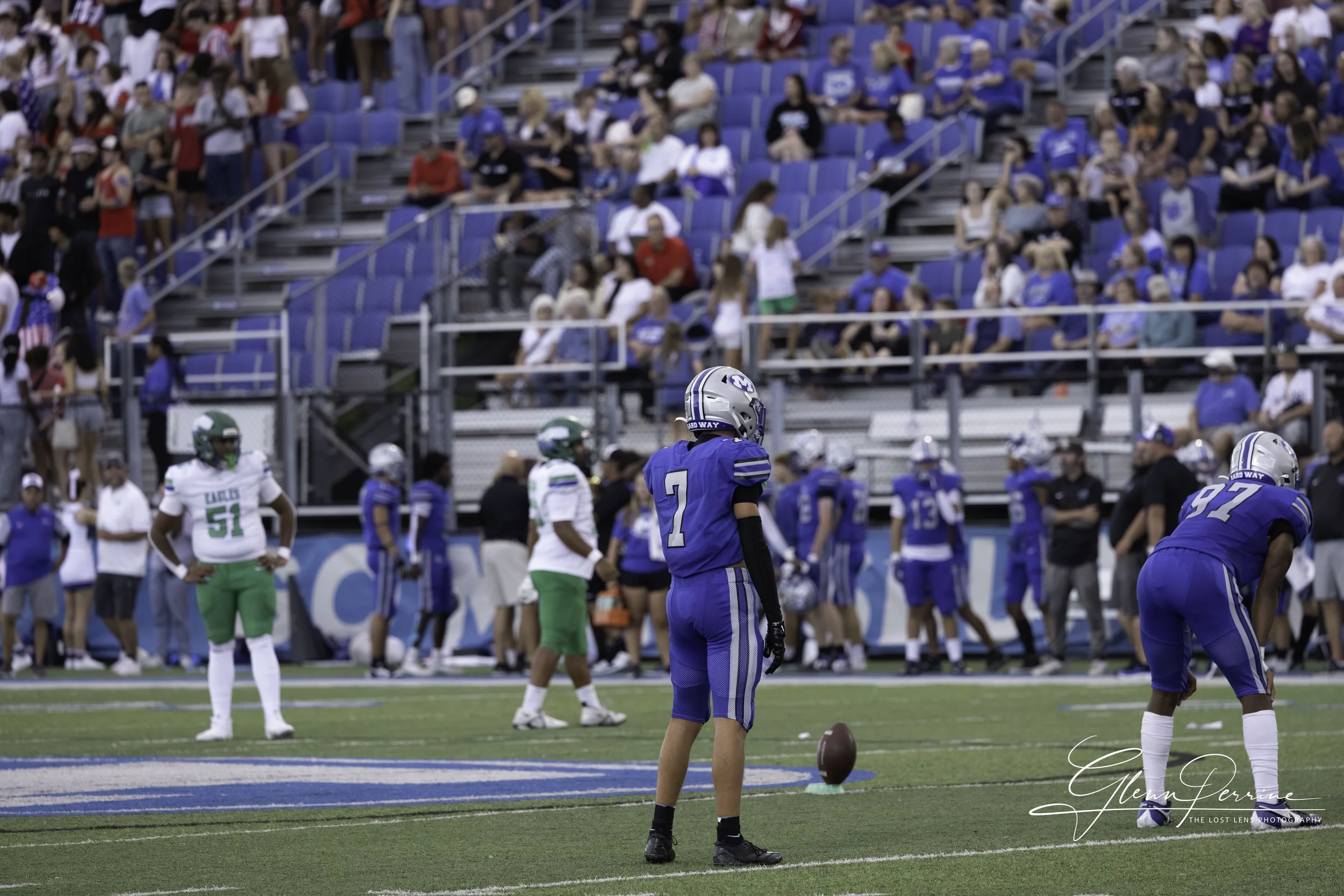 An American football game at a stadium with players on the field, some preparing for play, and spectators in the bleachers.