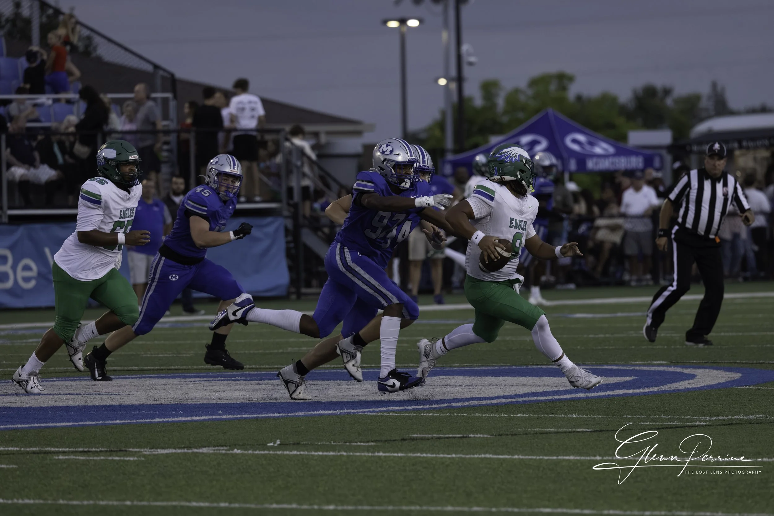 American football game with players in blue and green uniforms. Player in green with the ball tries to advance while players in blue chase him on the field. Crowd and a referee are visible in the background.