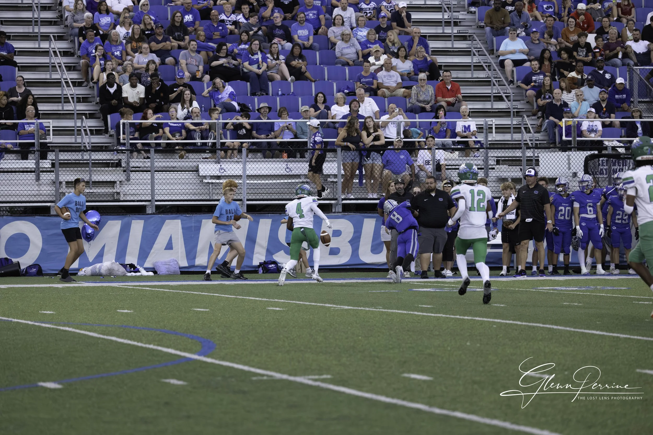 Pop Warner football game with players, coaches, and cheerleaders on the field, spectators in the stands, and advertising banners along the sideline.