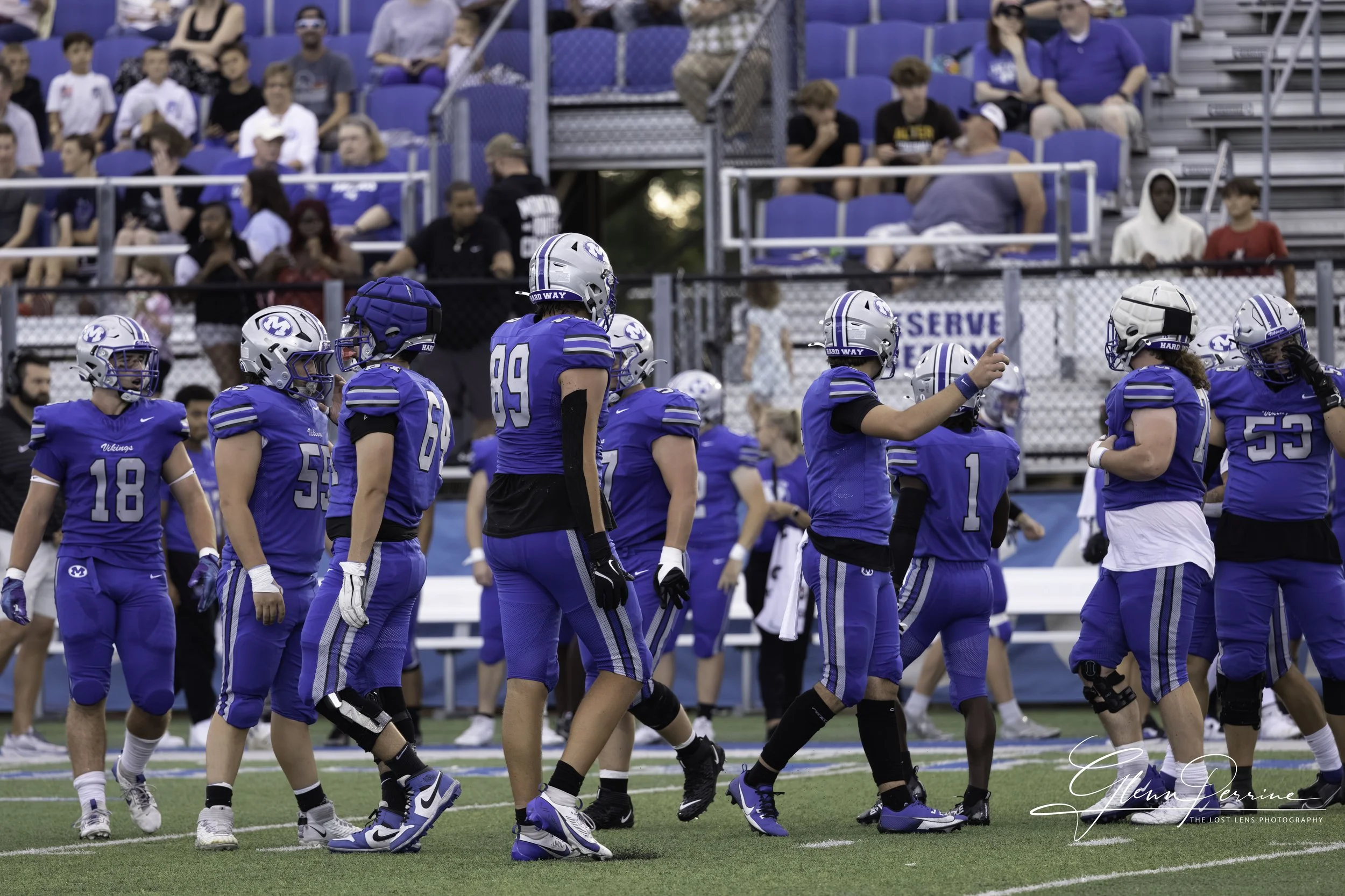 A group of football players in blue uniforms and helmets walking on the field, with spectators in the stands behind them.