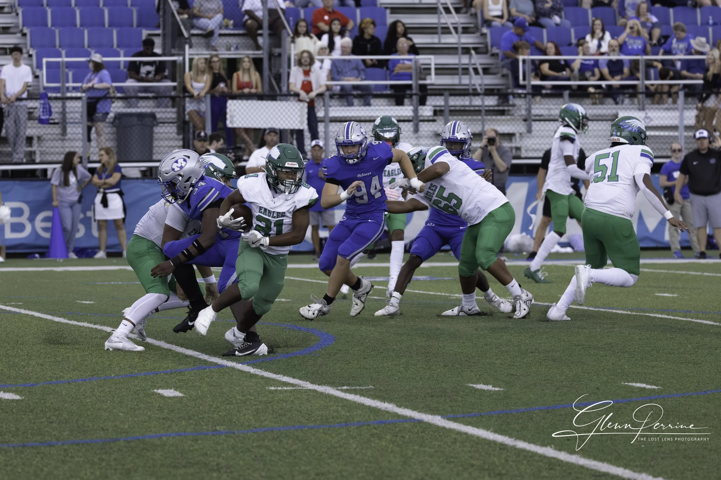 Football players in blue and white uniforms play a game on a field with spectators watching from the bleachers.
