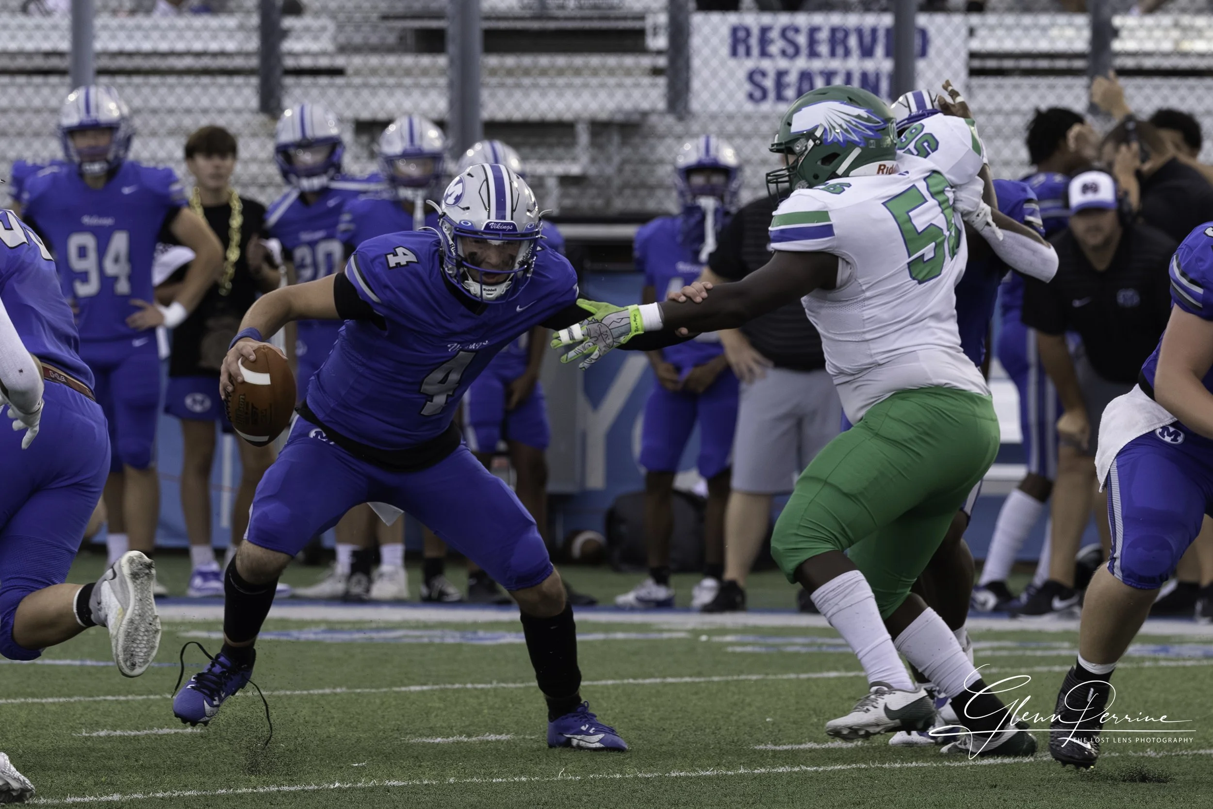 American football game with a player running with the football being tackled by an opponent, other players on the sideline observing, and a fence in the background.