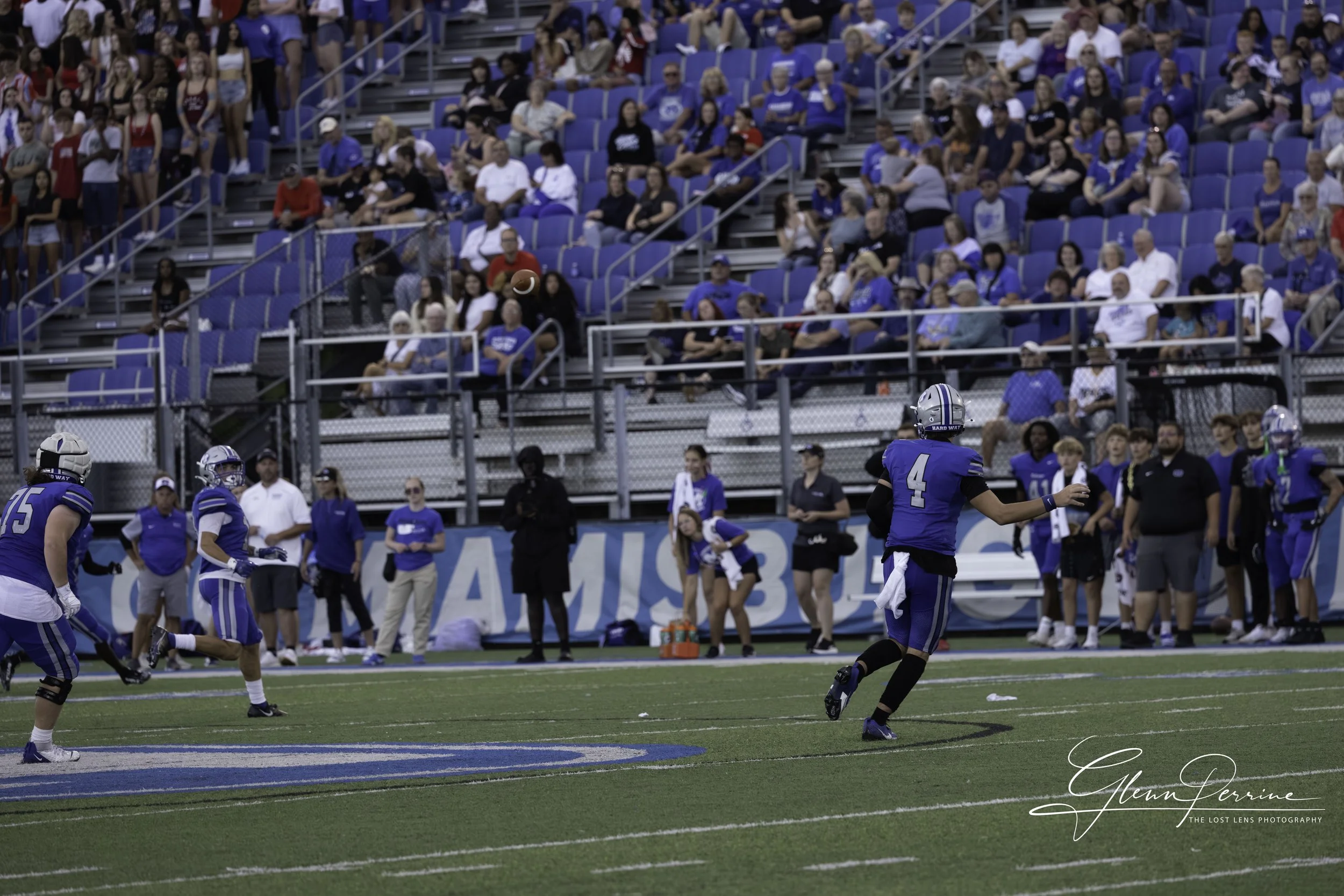 American football game with players in blue uniforms on the field, spectators in the stands watching, and a football in the air.