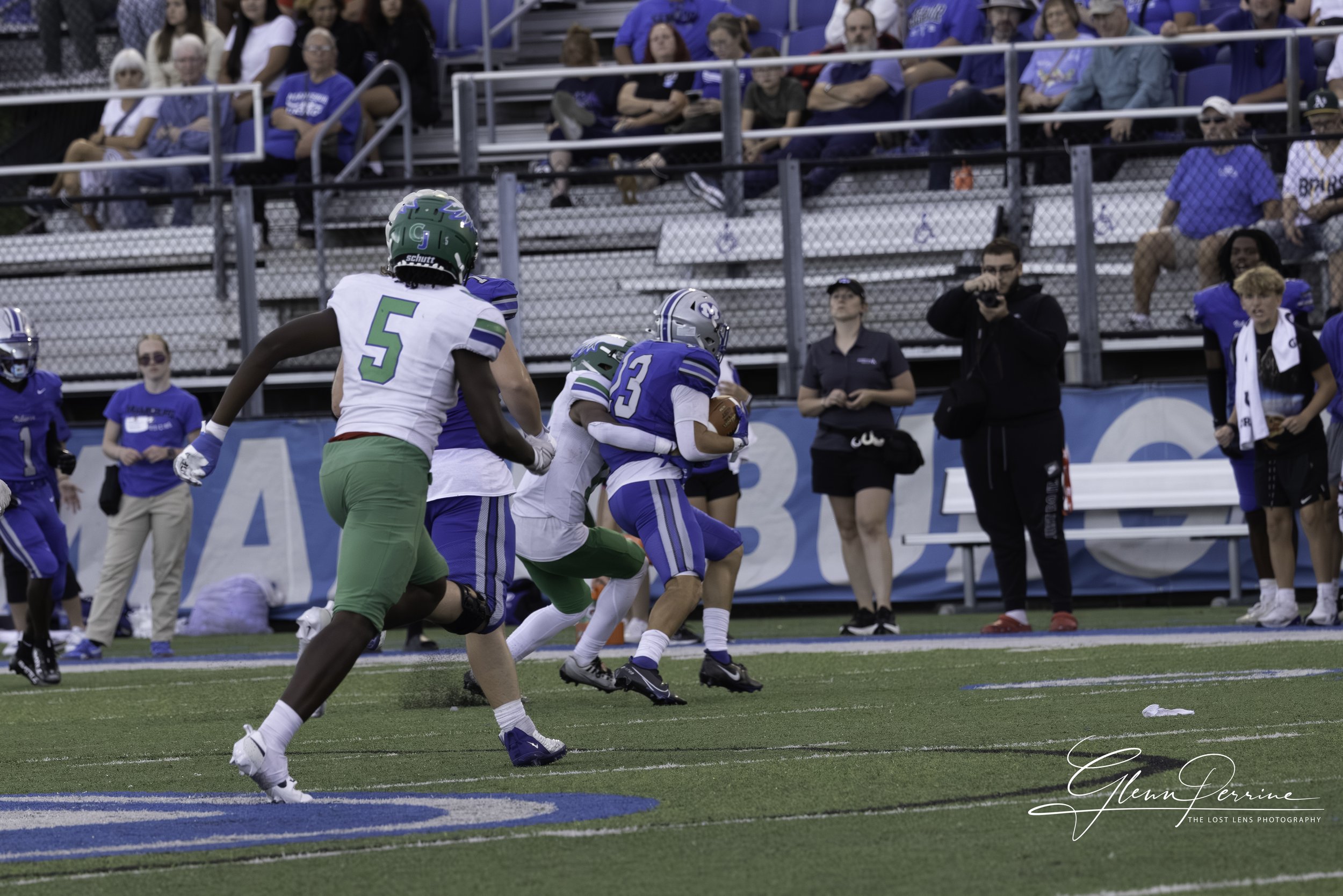 A football game where a player in a blue and white uniform is running with the ball while being chased by players in green and white uniforms on the field. Spectators are watching from the stands.