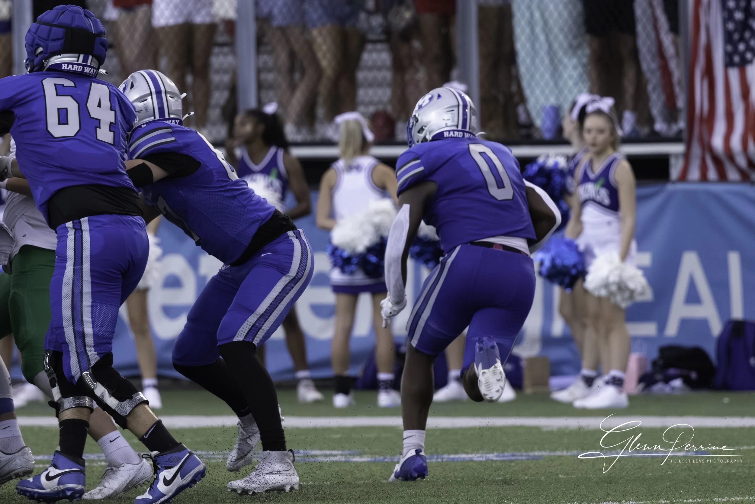 Football players in blue uniforms during a game, with cheerleaders watching in the background.