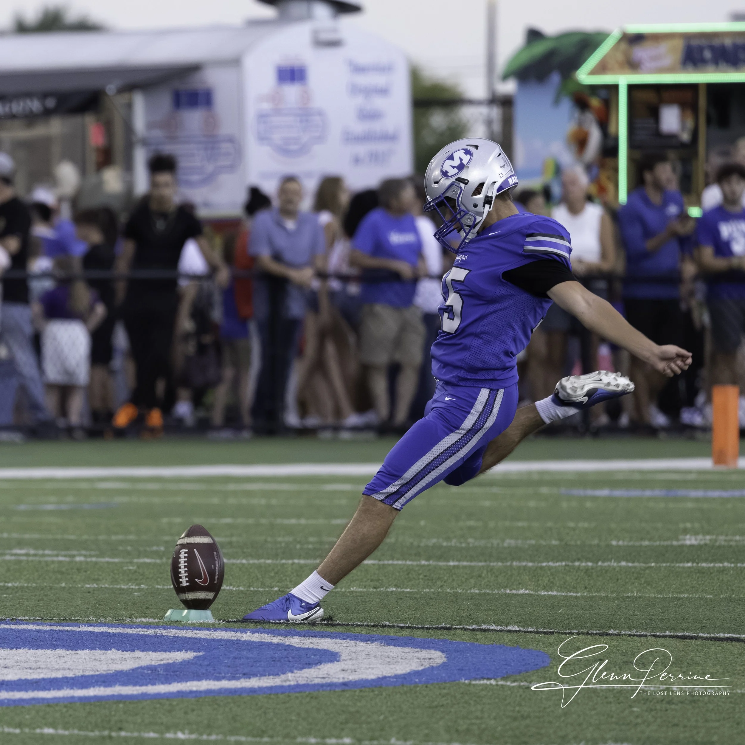 A football player in a blue uniform kicking a football on a field, with spectators in the background.
