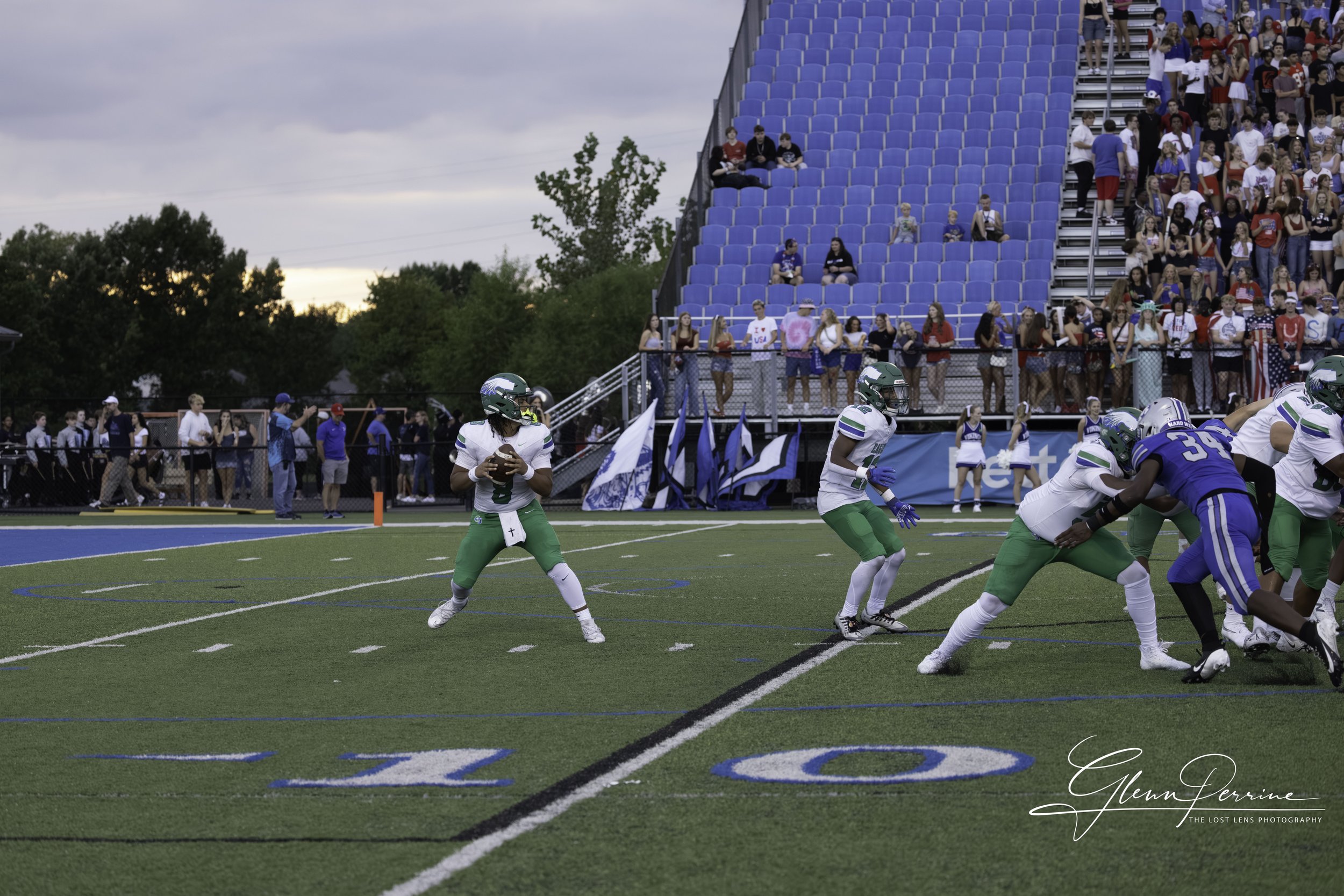 A football game in progress with players on the field, a quarterback preparing to throw the ball, and fans watching from the stands.