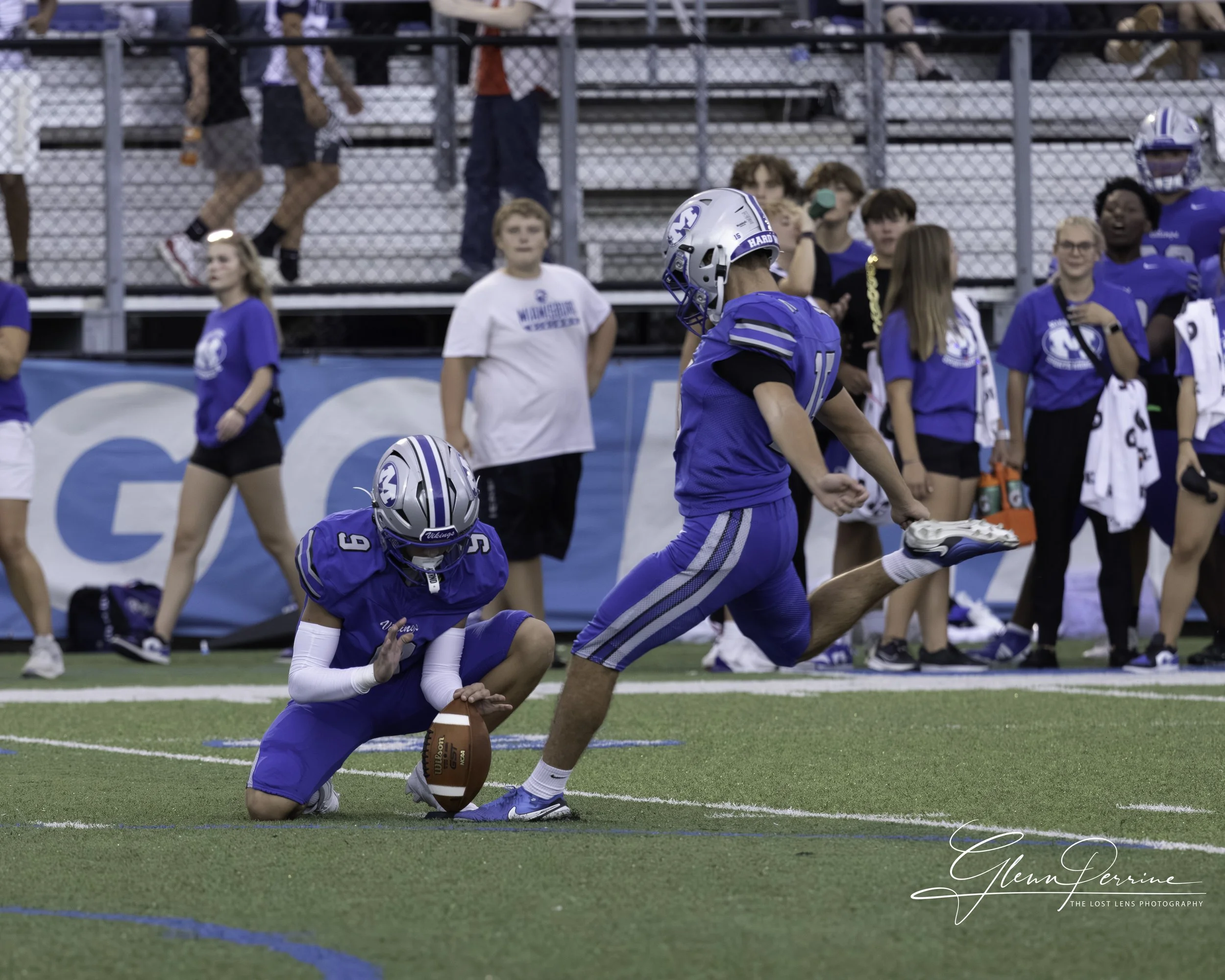 Two football players in blue uniforms and helmets on a football field, one kneeling with a football, the other running with a hand on the ground and holding a white towel, with spectators and team members in blue shirts on the sidelines.