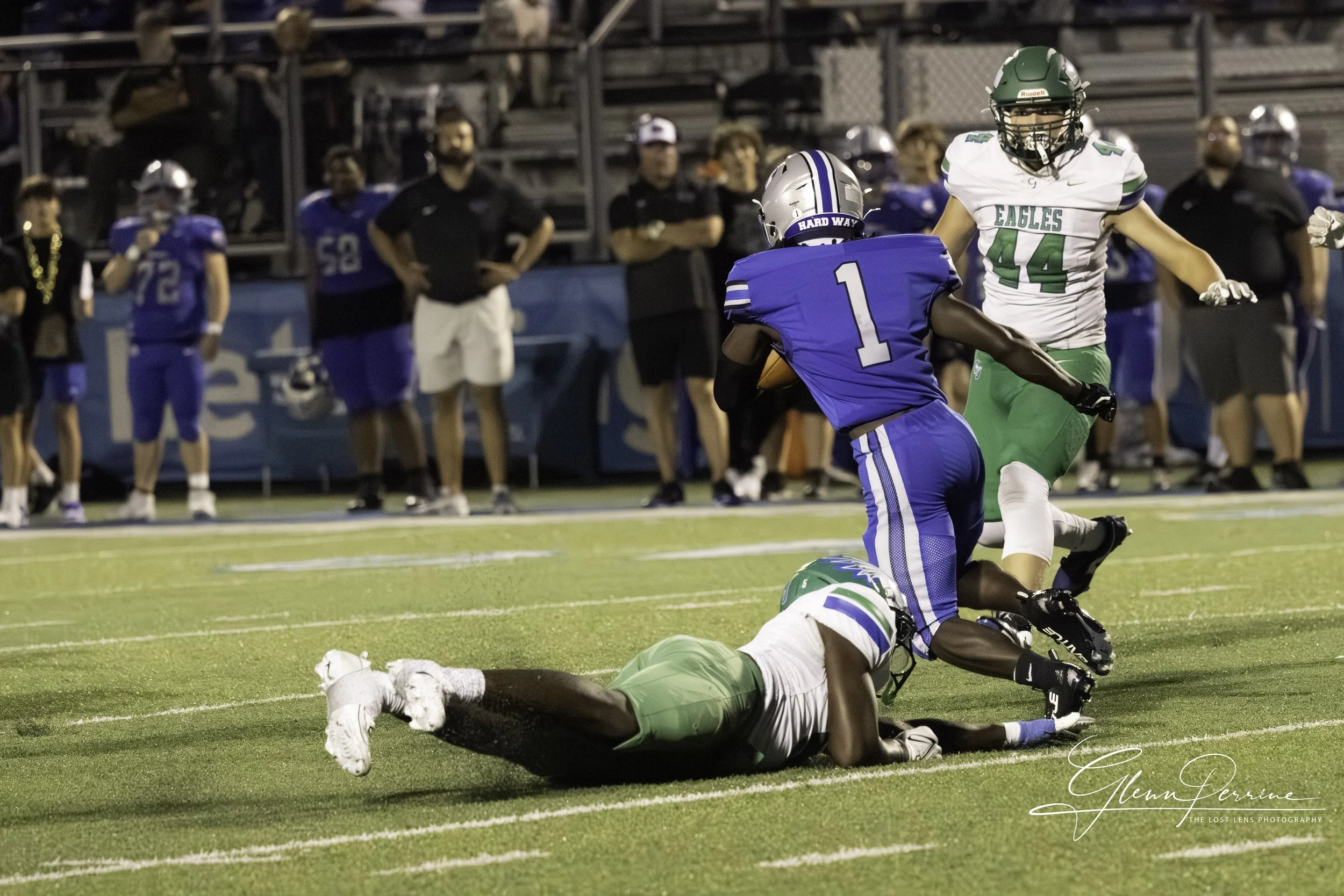 Football players during a game, with one player in a blue jersey carrying the ball and being tackled by a player in a green and white jersey on a turf field, with spectators and coaches on the sidelines.