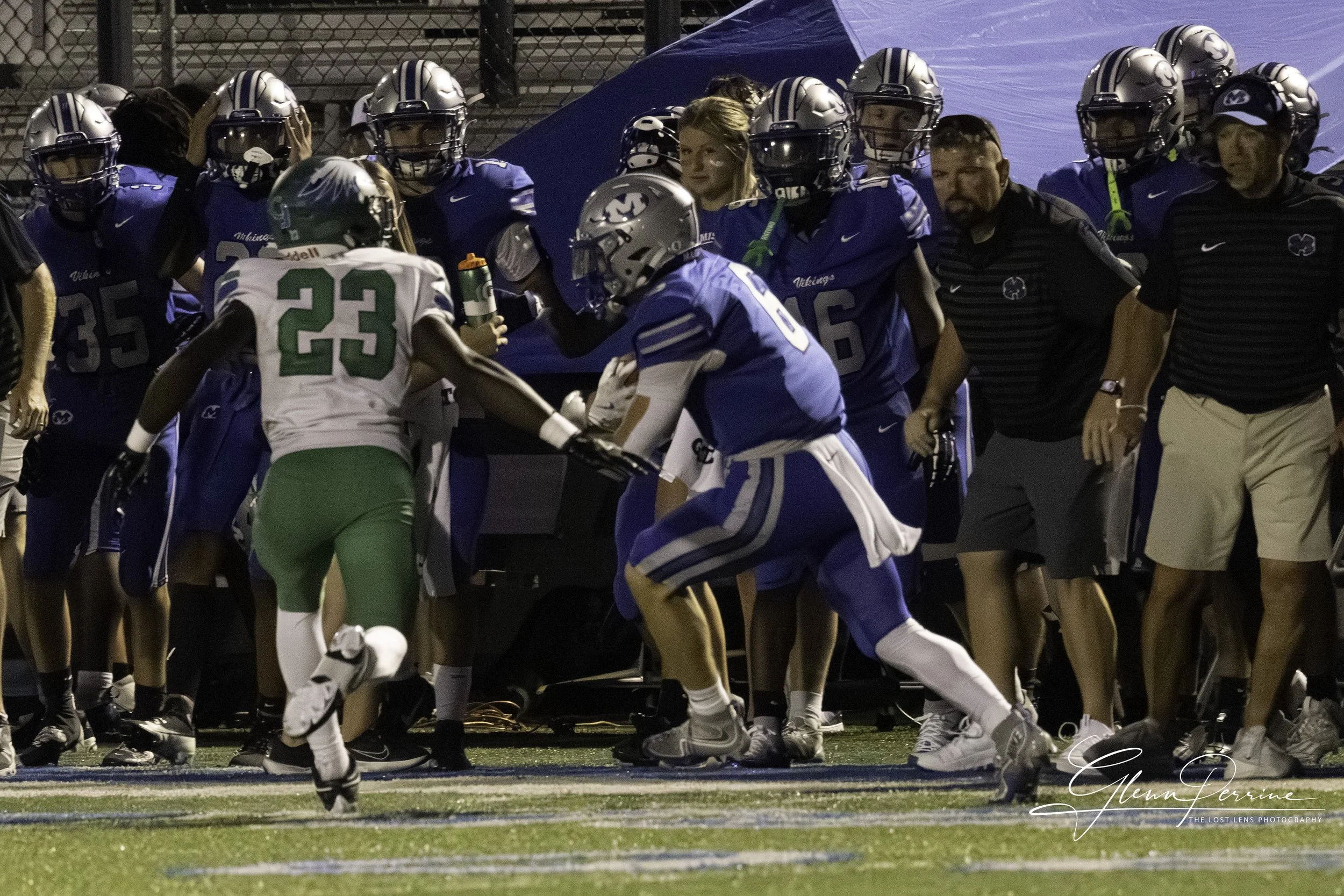 Football players in blue and white uniforms on the sideline, with two players in the foreground engaged in a tackle or block, one in a green and white uniform holding a water bottle, and coaches and team members watching from the sidelines.