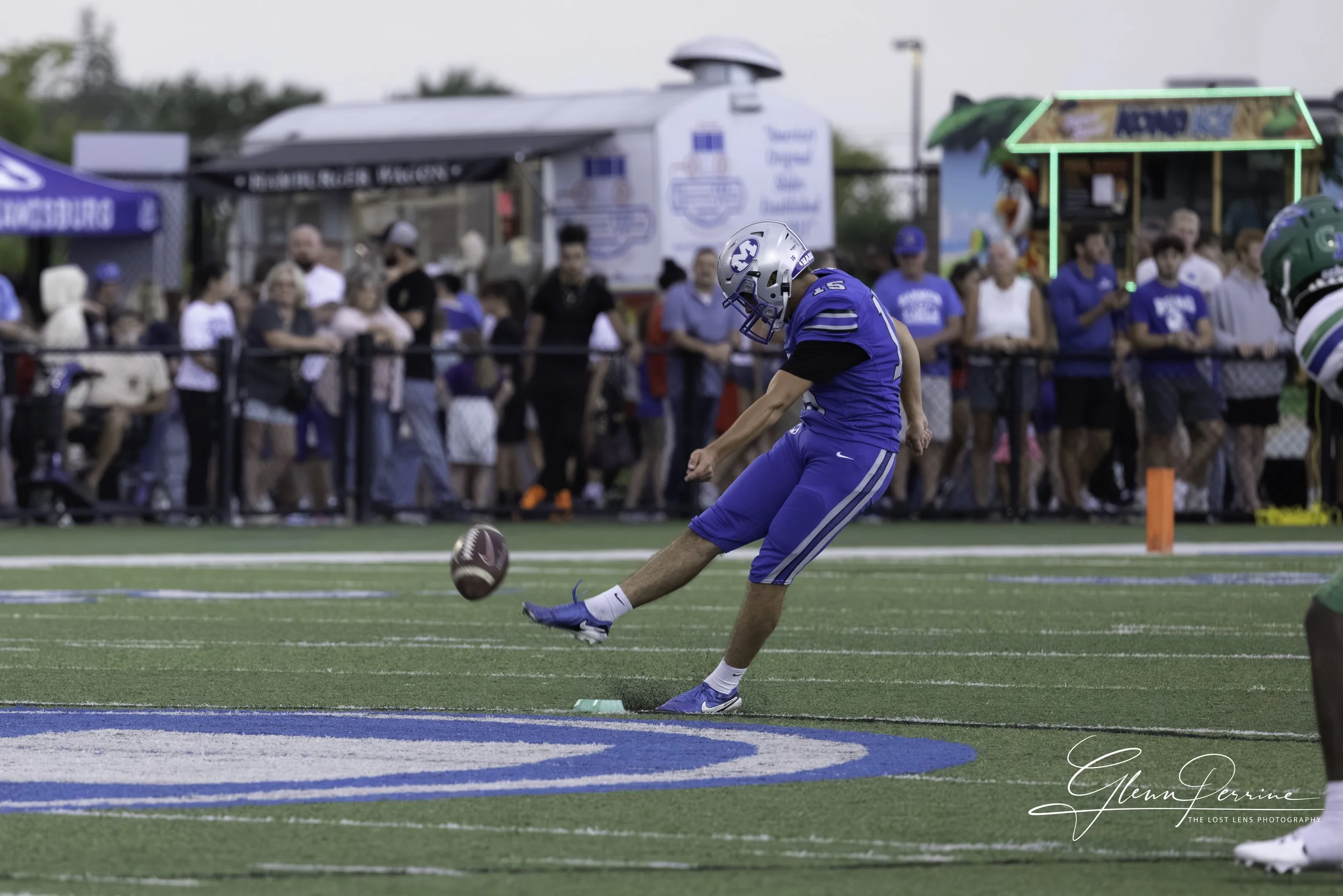 A football player in a blue uniform and helmet kicks a football on a field with spectators in the background.