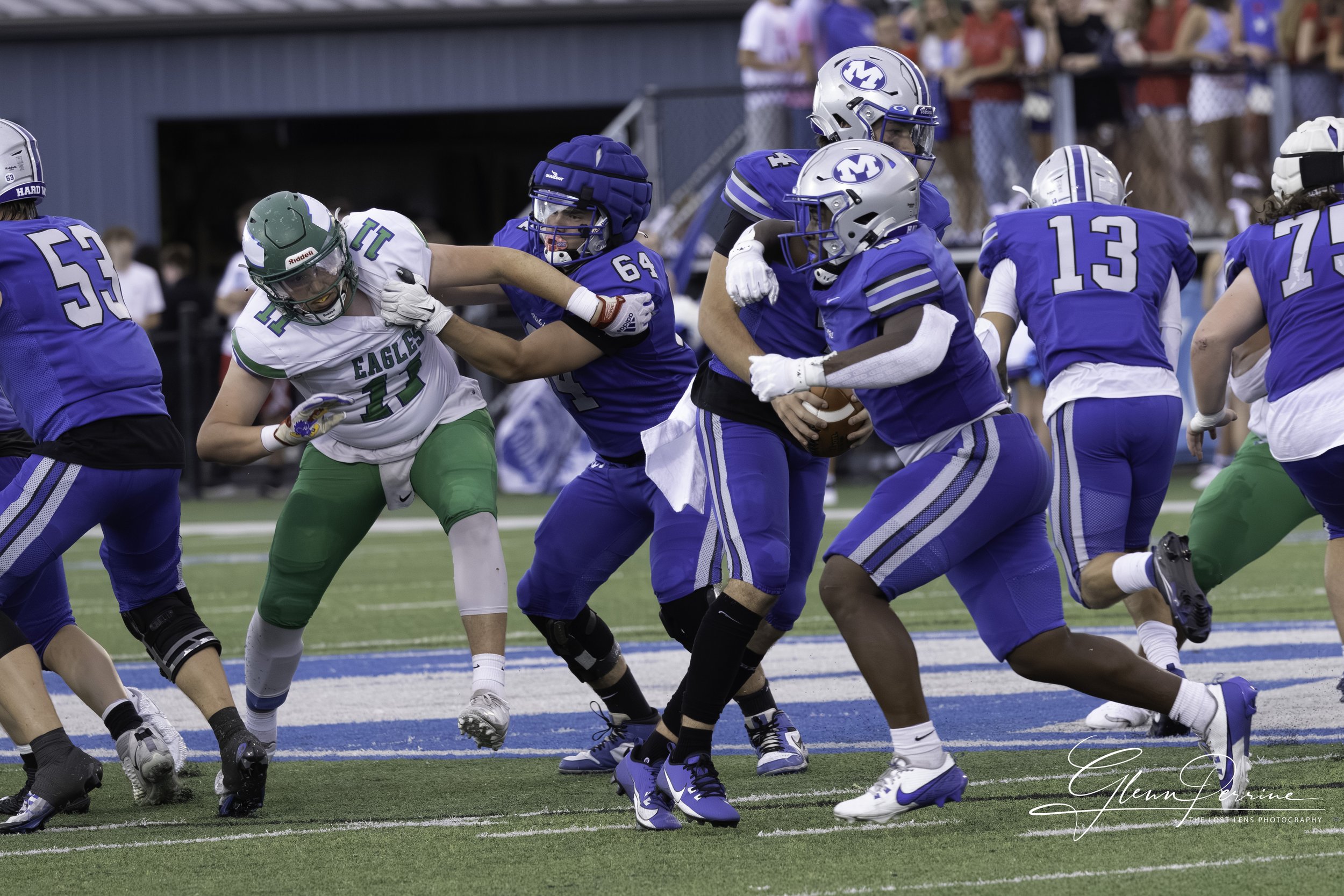 A football game with players in blue and green uniforms. The player in green is being tackled by a player in blue. The football is held by a player in blue. Spectators are visible in the background.